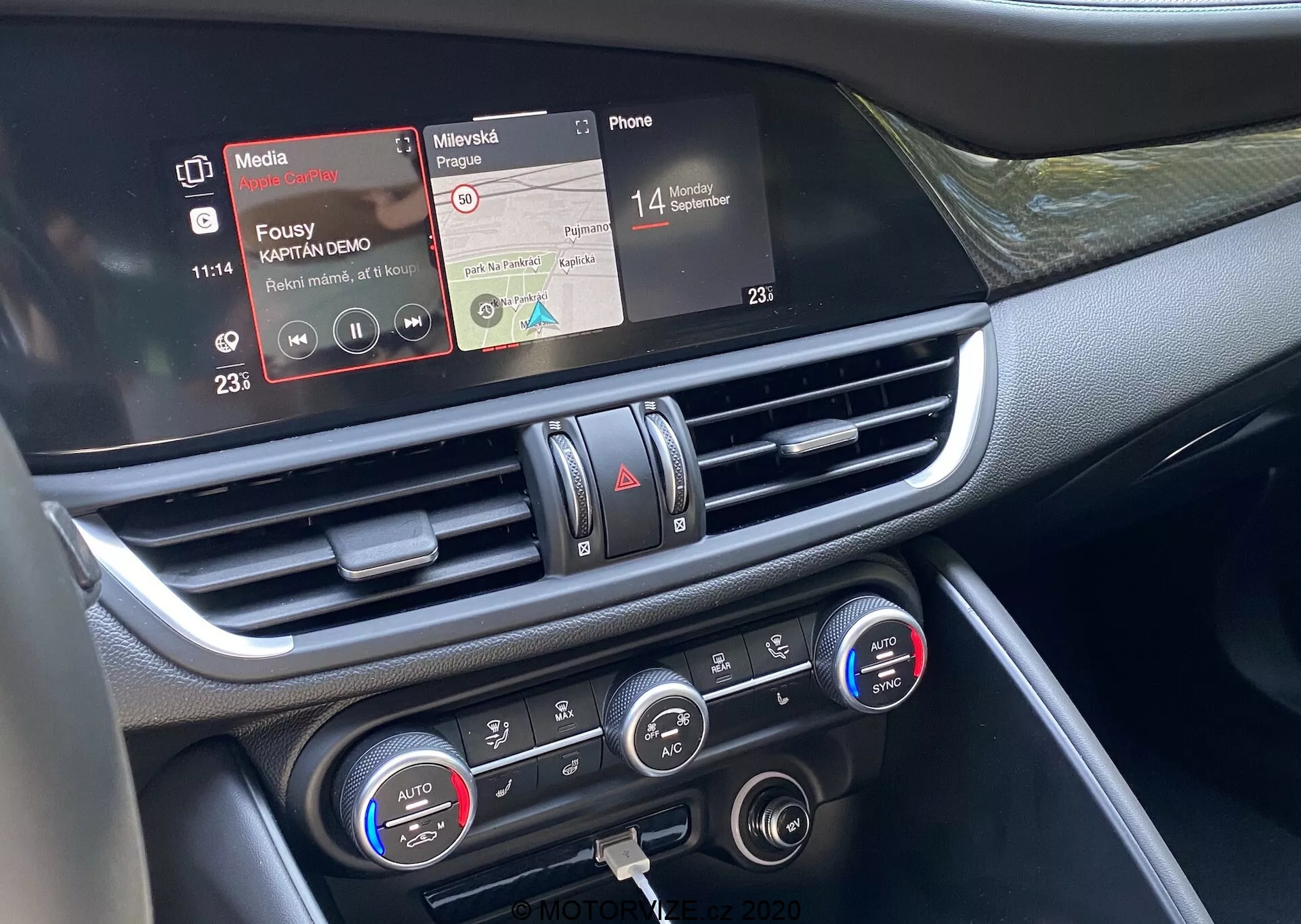 Detailný pohľad na centrálnu časť palubnej dosky Alfy Romeo Giulia 2016 z perspektívy predného spolujazdca. Stredne blízky záber zvýrazňuje dotykový infotainment displej s funkciami prehrávača médií a konektivity telefónu, centrálny výduch vzduchu s chrómovými akcentmi a ovládanie klimatizácie s nastaveniami dvojzónovej teploty a digitálnymi displejmi. Materiály palubnej dosky sa zdajú byť z plastov a kovových alebo chrómovaných prvkov, čo ukazuje pokročilú technológiu interiéru vozidla zameranú na konektivitu, prehrávanie médií a kontrolu klímy.