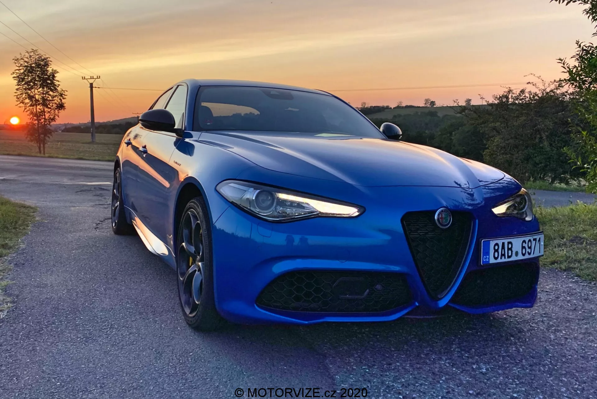 Modrý športový sedan Alfa-Romeo Giulia 2016 zachytený večer so západom slnka v pozadí, mierne naklonený smerom k fotoaparátu na dláždenej vidiecke ceste, predstavujúci charakteristickú prednú masku, agresívny dizajn LED predných svetiel, veľké viacramenné zliatinové kolesá a vysokovýkonný brzdový systém. Kovový lak, aerodynamický predný nárazník, tvarované línie kapoty a pokročilé senzory pre asistenčné systémy vodiča zdôrazňujú športovú estetiku a moderné technológie vozidla. Prítomný je vodotlač 'MOTORVIZE.cz 2020'.