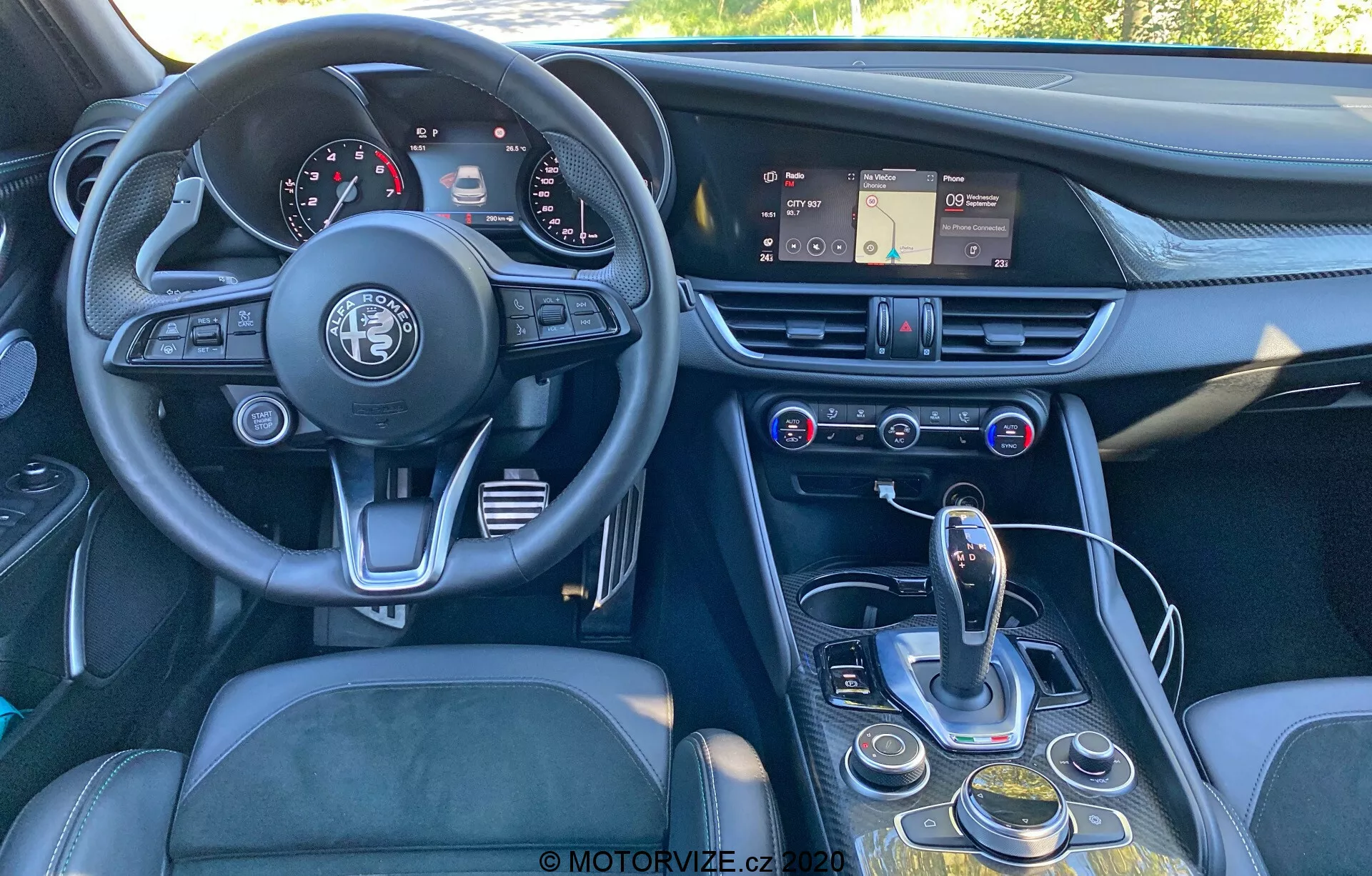 Pohľad na interiér Alfa Romeo Giulia z roku 2016 zo slabšie mimo stred, zadný na predný uhol, zobrazujúci prístrojovú dosku, volant, stredovú konzolu a časť vodičovho sedadla. Obrázok zvýrazňuje trojramenný volant s emblémom Alfa Romeo, prístrojovú dosku s analógovými ciferníkmi a digitálnymi displejmi, palubnú dosku s dotykovým infotainmentovým systémom, vetracie otvory a ovládacie prvky klimatizácie a multimédií. Stredová konzola obsahuje automatický volič prevodovky, otočný číselník, tlačidlá pre navigáciu infotainmentu a nastavenia vozidla a možný volič režimu. Dizajn kladie dôraz na ergonómiu a prístupnosť, s materiálmi ako koža, kefovaný kov a vysoko kvalitný plast, aby navodil pocit luxusu.