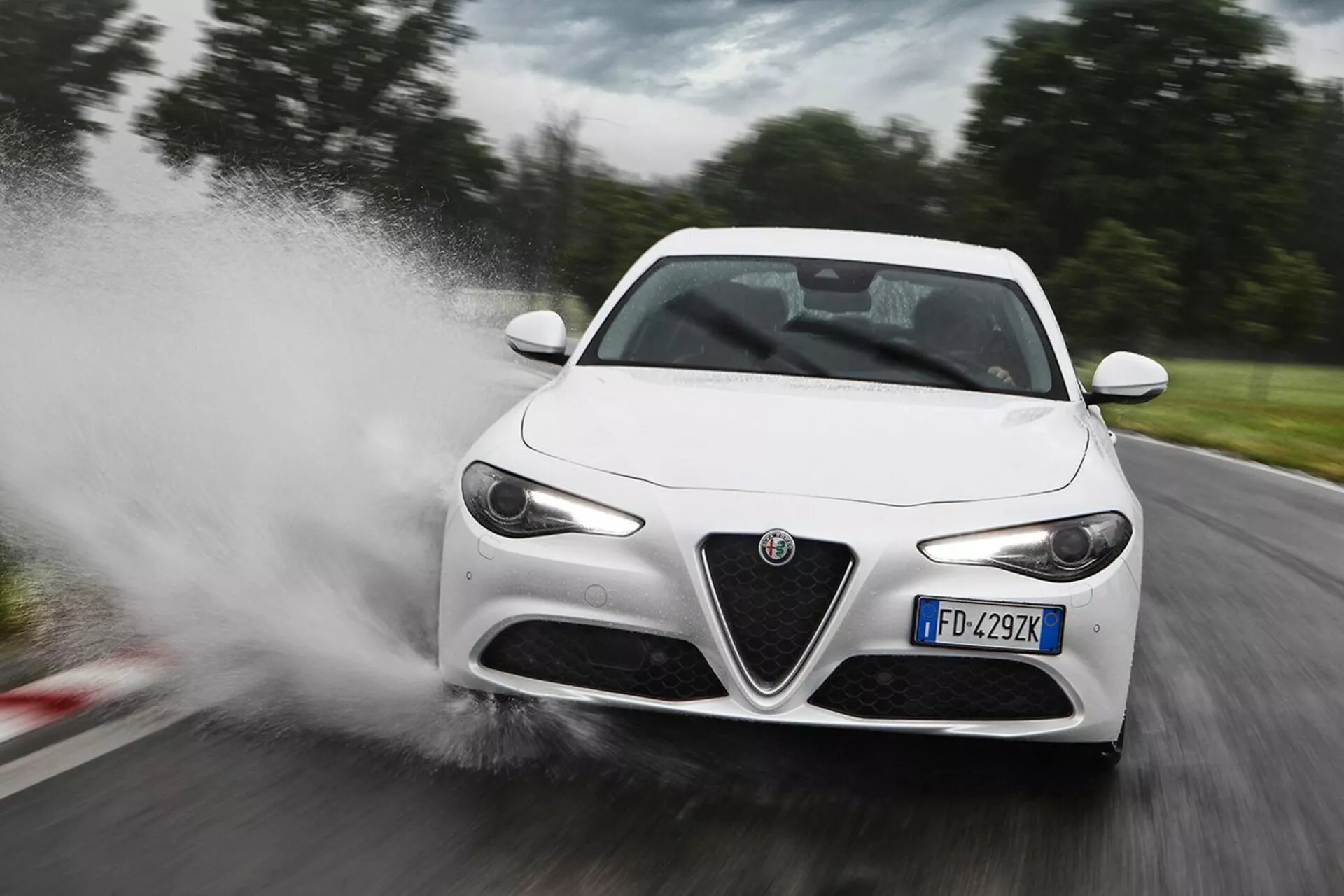 Biely športový automobil Alfa Romeo Giulia z roku 2016, ktorý prechádza vysokou rýchlosťou cez vodu, spôsobujúc rozstrek, v trojštvrťovom prednom zornom uhle zdôrazňujúcom jeho LED svetlomety, charakteristickú prednú mriežku, elegantné bočné zrkadlá a aerodynamické línie karosérie