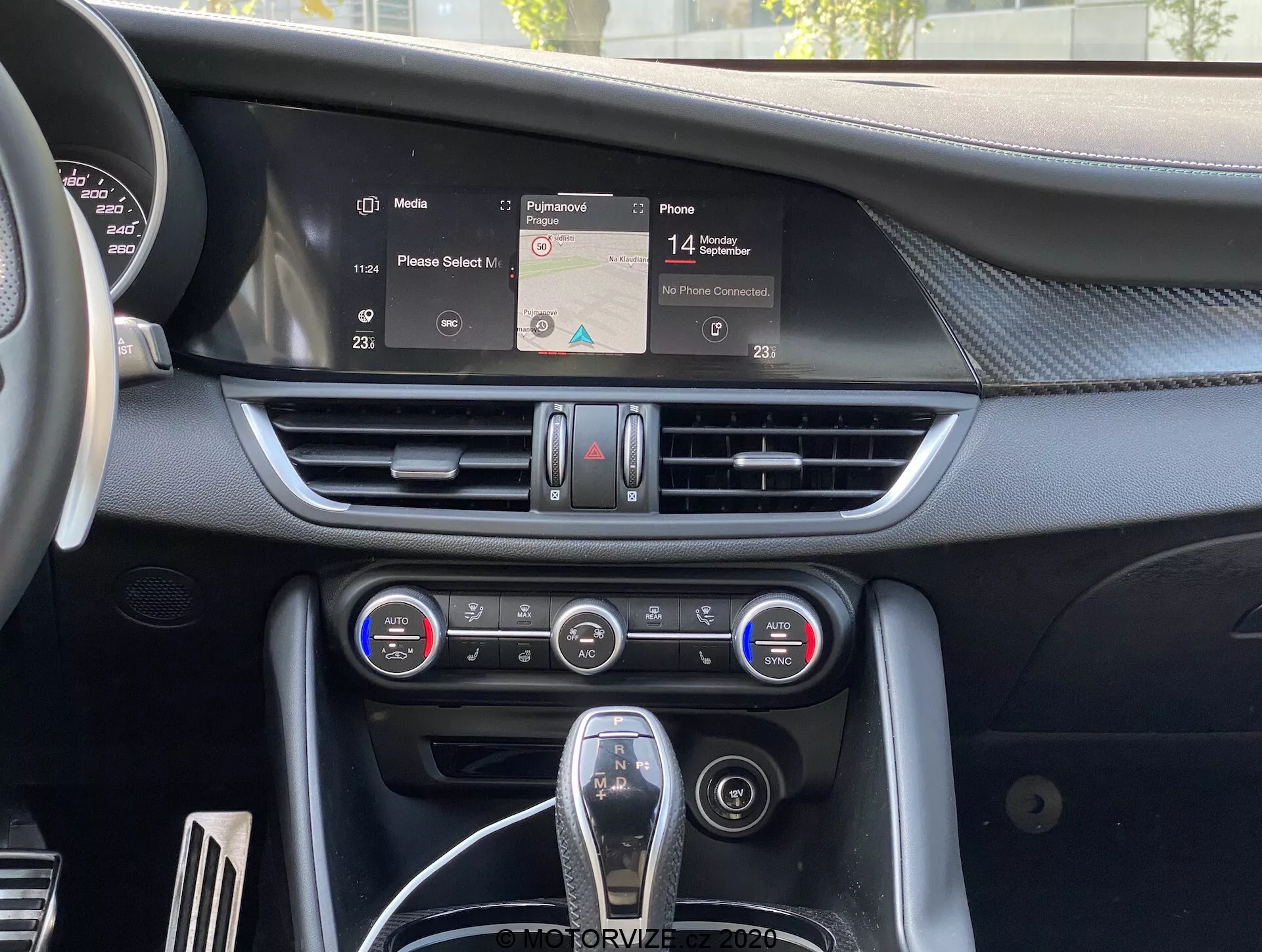 Pohľad do interiéru Alfa-Romeo Giulia 2016 so zameraním na stredovú konzolu so zobrazenou navigačnou mapou na infotainment displeji, horizontálne vetracie otvory, dvojzónový klimatizačný panel s tlačidlami a gombíkmi a čiastočne viditeľným voličom prevodov s koženým poťahom a chrómovými akcentmi, zobrazujúci modernú ergonomiu a technológie interiéru auta.