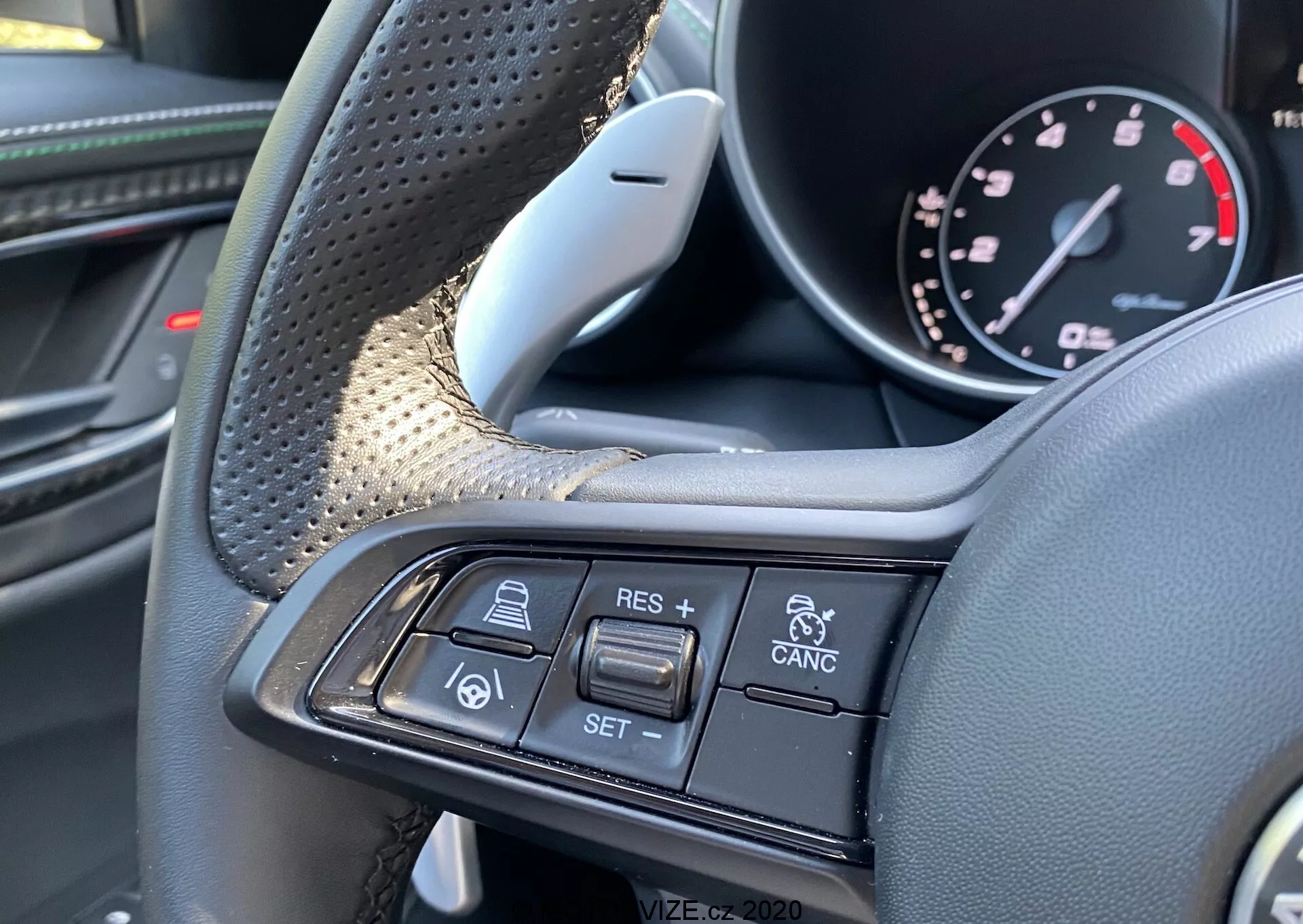 Detailný pohľad na interiér Alfa-Romeo Giulia 2016, zobrazujúci volant a jeho multifunkčné ovládacie prvky z pohľadu vodiča. Na ľavej strane sú viditeľné textúrované úchopy a luxusné materiály ako koža a možno aj karbónové vlákna. Pravé rameno volantu obsahuje tlačidlá tempomatu a adaptívneho tempomatu, vrátane + RES, - SET, CANCEL, ikony auto-nasledujúce-auto a varovania pred kolíziou vozidiel. Obrázok sa sústredí výhradne na tieto ovládacie prvky, zdôrazňujúc prémiový dizajn a pokročilé asistenčné funkcie pre vodiča.