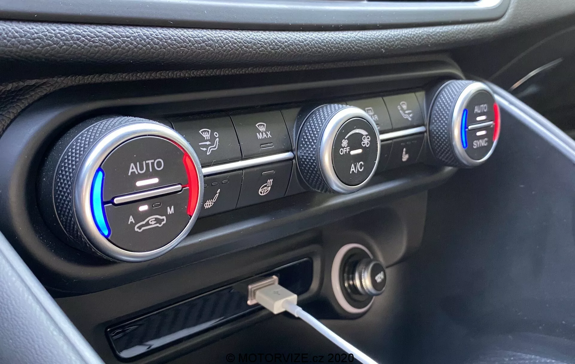 Detailný záber na klimatizačný systém na prístrojovej doske modelu Alfa Romeo Giulia 2016 s troma kruhovými ovládačmi. Ľavý ovládač ukazuje nastavenie teploty s modrým a červeným pásom a nastavením 'AUTO', čiastočne osvetleným na červeno. Stredný ovládač zobrazuje nastavenie rýchlosti ventilátora, zvýraznené na nižšej rýchlosti. Pravý ovládač zobrazuje rôzne režimy rozvodu vzduchu s tlačidlami 'AC' a 'SYNC'. Pod ovládačmi je 12-voltová zásuvka a USB port. Prístrojová doska má čierne prevedenie so strieborným akcentom okolo ovládania klimatizácie, zachytené z vyvýšeného uhla.