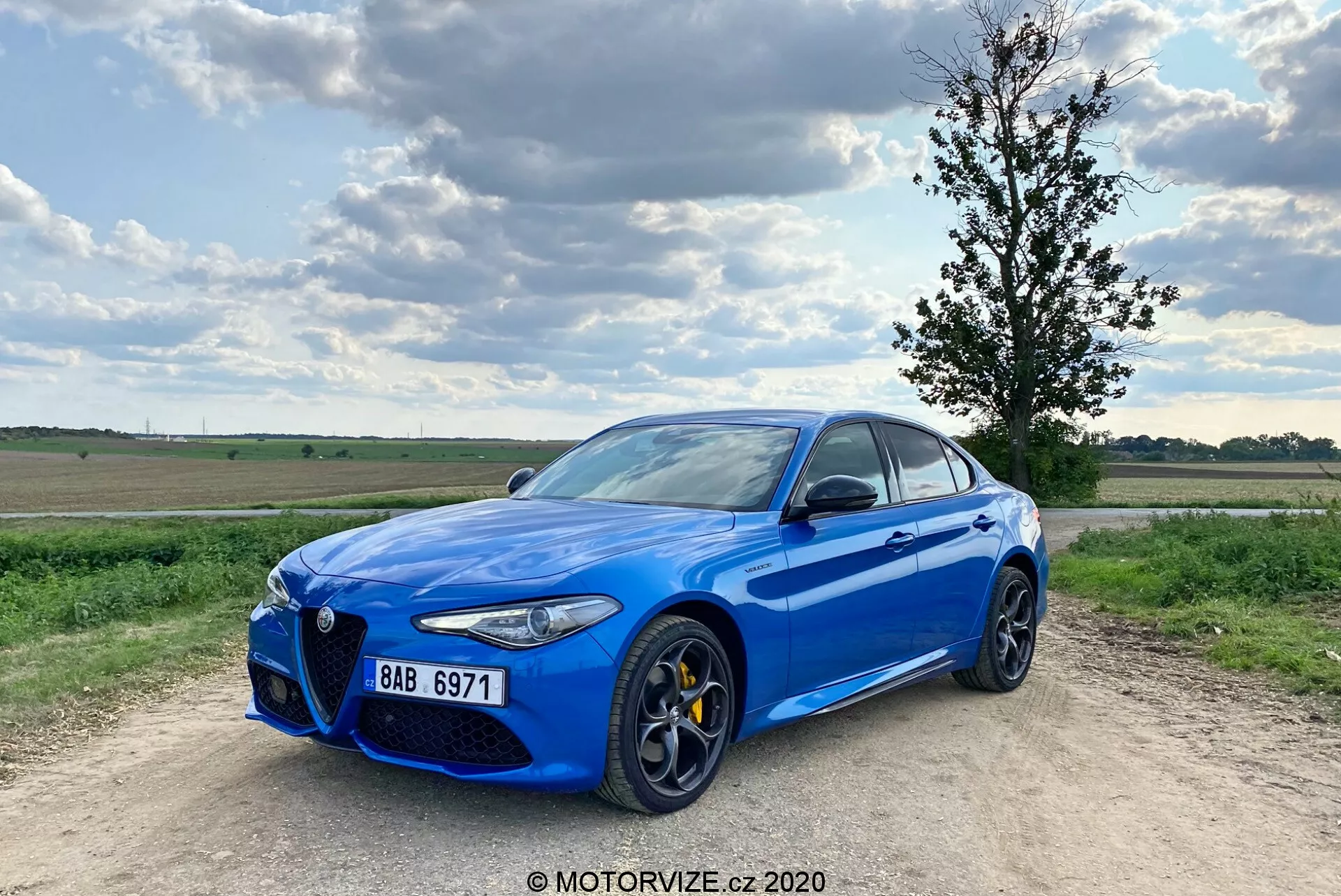 Obrázok jasne modrého športového sedanu Alfa Romeo Giulia 2016 zaparkovaného na štrkovej ceste s čiastočne oblačnou modrou oblohou v pozadí. Auto je zachytené z trojštvrťového predného uhla, ukazujúc jeho charakteristický predný mriežkový dizajn 'Trilobo', žlté brzdové strmene, veľké viacramenné zliatinové kolesá, elegantné LED svetlá a chrómové okenné obruče. Vozidlo je mierne vľavo od stredu s predným ľavým kolesom natočeným von, čo mu dodáva dynamický postoj.