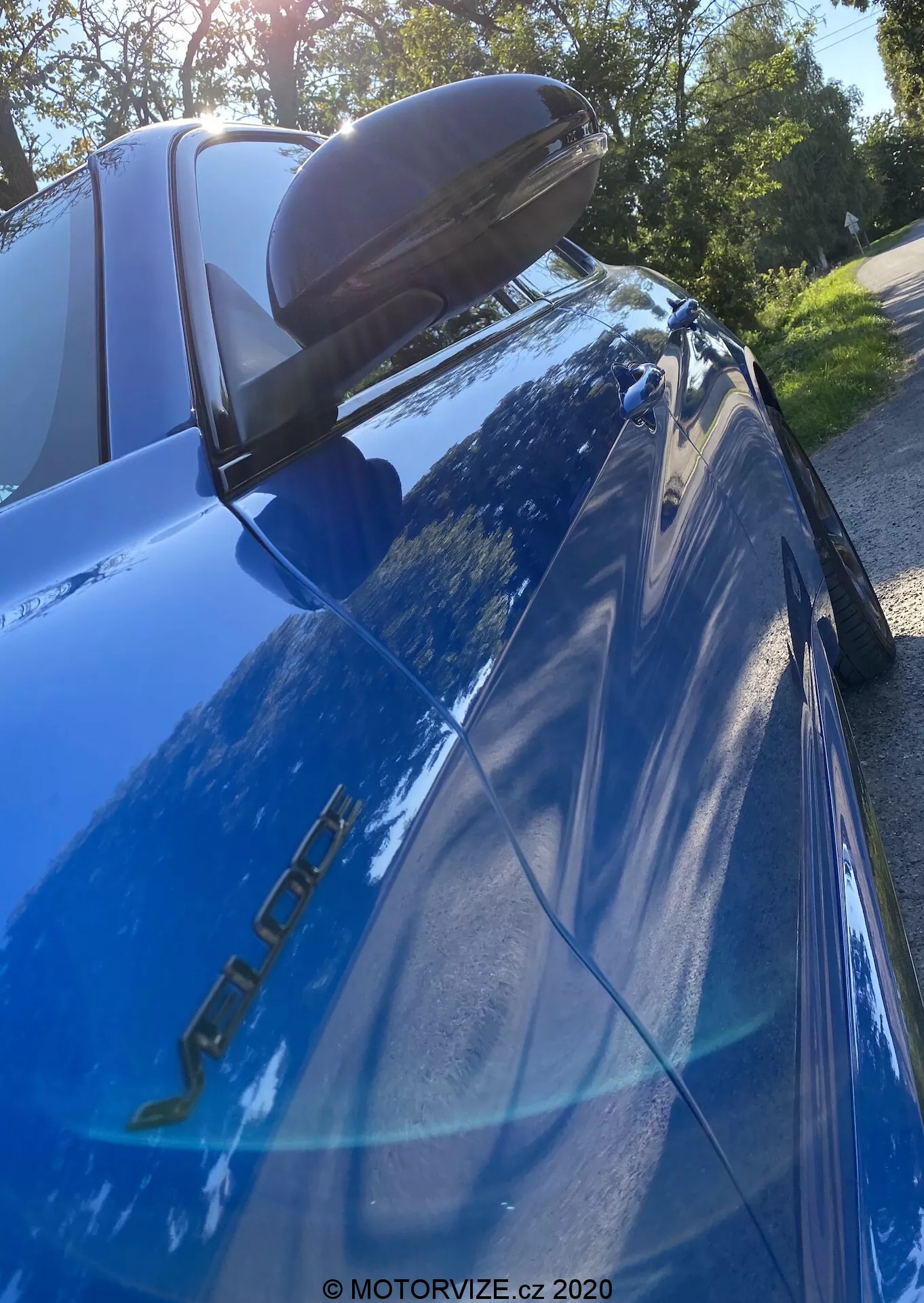 Detailný obrázok modrého Alfa Romeo Giulia (model 2016) zhotovený z nízkeho a nahor smerujúceho uhla so zameraním na zadnú časť. Viditeľné sú modelové znaky na strane vodiča, reflexný modrý lak, ľavé zadné LED svetlo, čierny zadný spojler, klesajúce zadné okno s tretím brzdovým svetlom, odraz stromov a jasnej oblohy a bočné zrkadlo s integrovaným smerovkami.