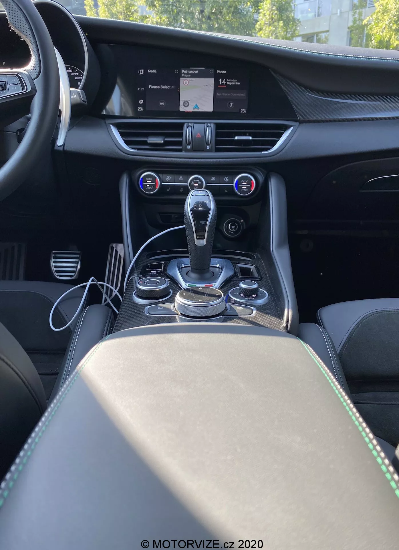 Interiér Alfa Romeo Giulia 2016 zobrazený zozadu centrálnou konzolou, zobrazujúci stredový panel, tunel prevodovky a časť sedadla spolujazdca. Obrázok poskytuje čelnú perspektívu z pohľadu zadného pasažiera, zdôrazňujúc modernú automatickú radiacu páku obklopenú kovovými ovládacími gombíkmi a tlačidlami pre infotainment systém a jazdné módy. Nad oblasťou radiacej páky sa nachádza dvojzónová automatická klimatizácia s digitálnym displejom ukazujúcim nastavenia teploty a tlačidlami na kontrolu distribúcie vzduchu a rýchlosti ventilátora. Veľká obrazovka infotainment systému na vrchole stredového panelu zobrazuje mapu navigačného systému. Palubná doska je vybavená detailmi šitia a kovovým nádychom, ktoré naznačujú prémiové materiály. Opierka rúk a úložný priestor s dvoma držiakmi na poháre sú viditeľné za radiacou pákou, čo zvyšuje luxusný a technologicky orientovaný dizajn kabíny.