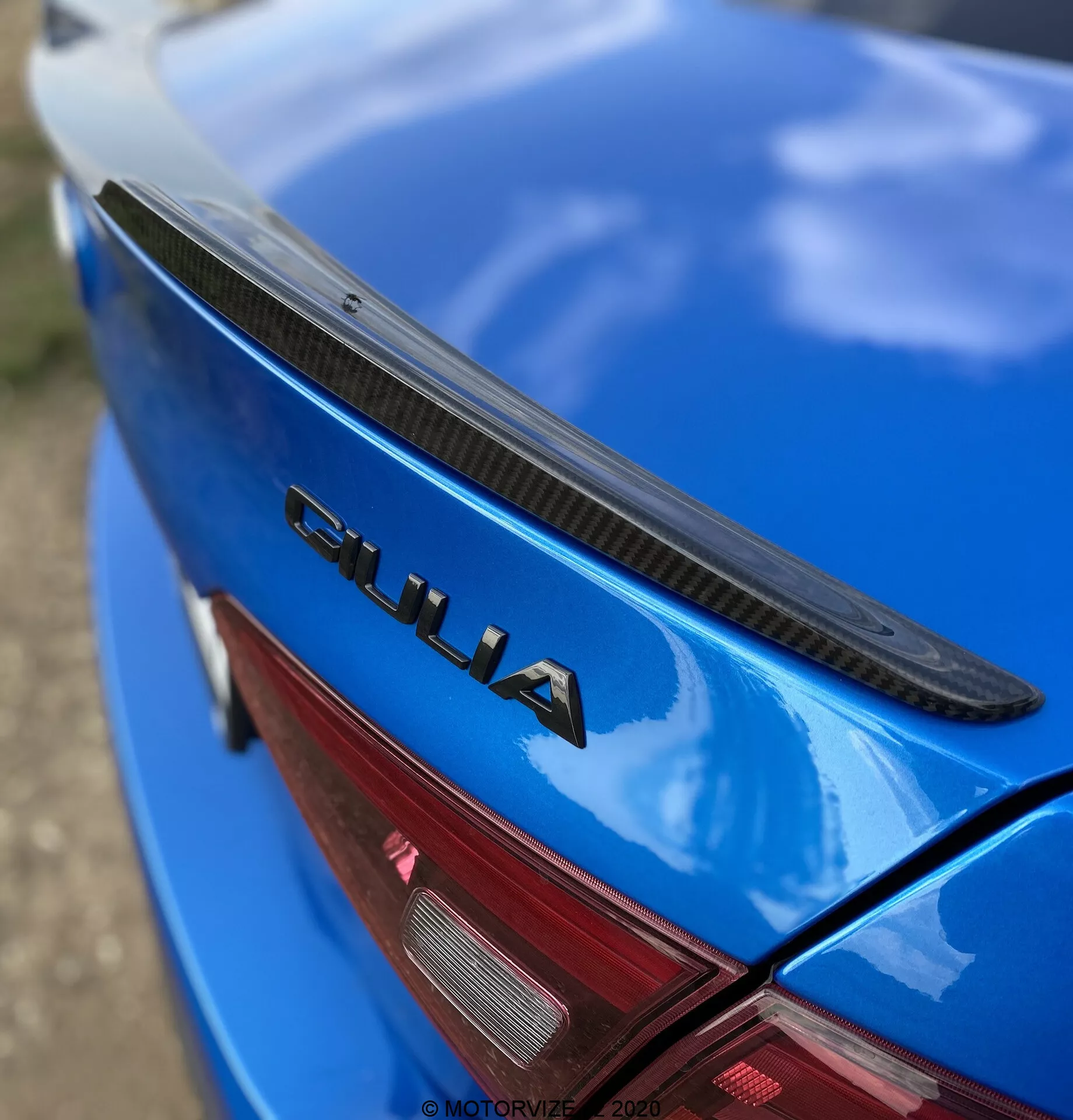 Detailný šikmý pohľad na zadnú časť modrého Alfa Romeo Giulia 2016. Fotografia zachytáva viečko kufra s čiernym spojlerom, lesklú modrú farbu a výrazným chrómovým znakom 'GIULIA'. Viditeľná je tiež časť červeného LED zadného svetla. Znak 'GIULIA' je ostro zameraný, zatiaľ čo spojler, povrch auta a zadné svetlo sa postupne rozostrujú, čím zdôrazňujú branding a dizajnové prvky auta.