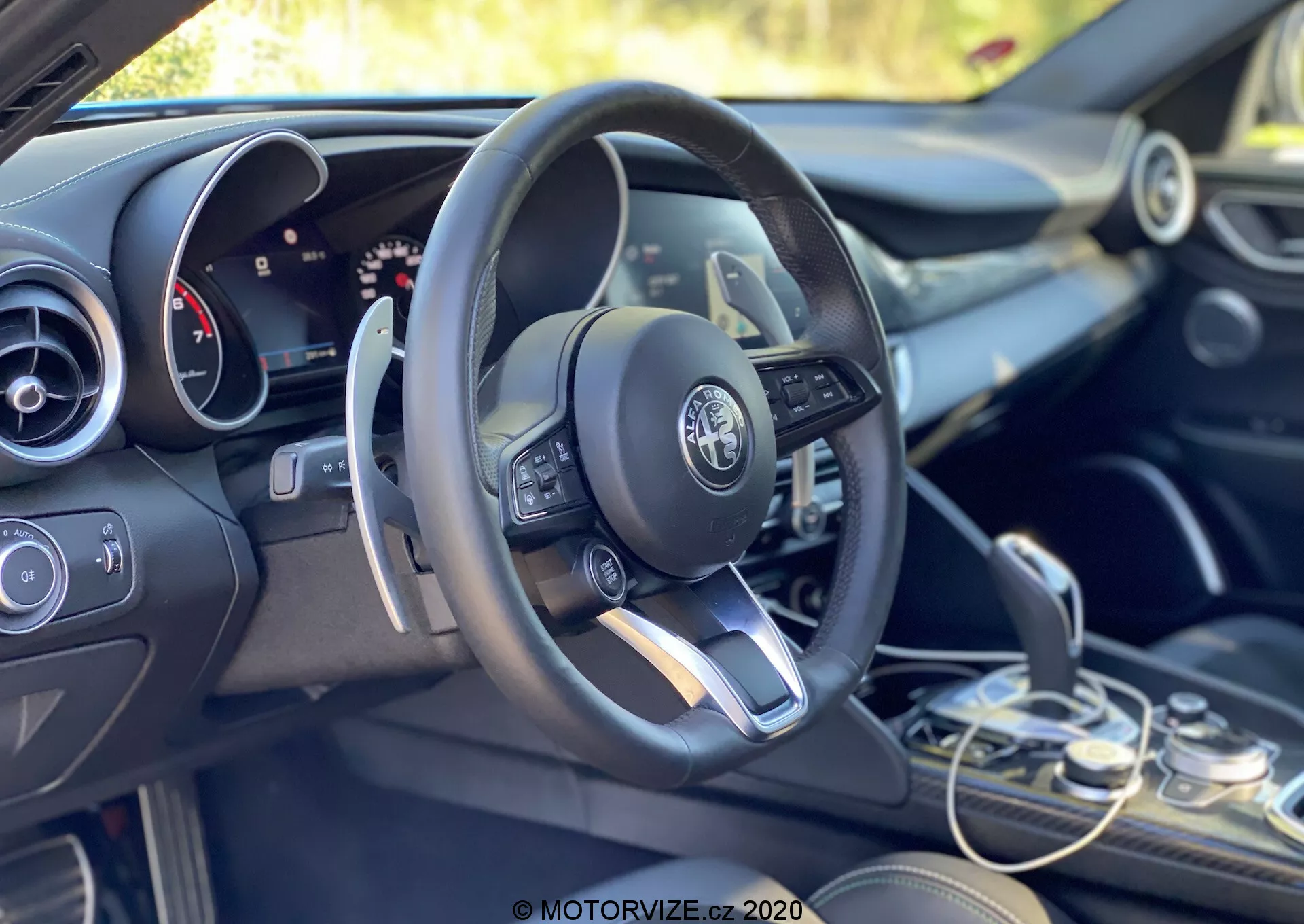 Pohľad na interiér vozidla Alfa Romeo Giulia 2016 zameraný na oblasť vodiča a prístrojovú dosku z ľavej strany, mierne zozadu. Konfigurácia pre ľavostranné riadenie zobrazuje moderný multifunkčný volant s namontovanými ovládacími prvkami, vrátane tlačidiel na nastavenie hlasitosti, prístup do menu a pravdepodobne aj tempomat. Logo Alfa Romeo je jasne viditeľné v strede volantu. Za volantom je čiastočne viditeľný kombinovaný analógový a digitálny panel, ktorý naznačuje konfigurovateľný informačný systém pre vodiča. Napravo od volantu je viditeľný horný okraj infotainment displeja, pod ktorým sú systémy klimatizácie a funkčné prepínače. Volič prevodovky, umiestnený v pravom dolnom rohu, ukazuje elegantný dizajn automatickej prevodovky. Vyhotovenie interiéru kombinuje tmavé tóny s kovovými akcentmi, čo odzrkadľuje prémiový dizajn vozidla. Kabína je zaliata denným svetlom, zelené pozadie naznačuje vonkajšie alebo vegetačné prostredie.