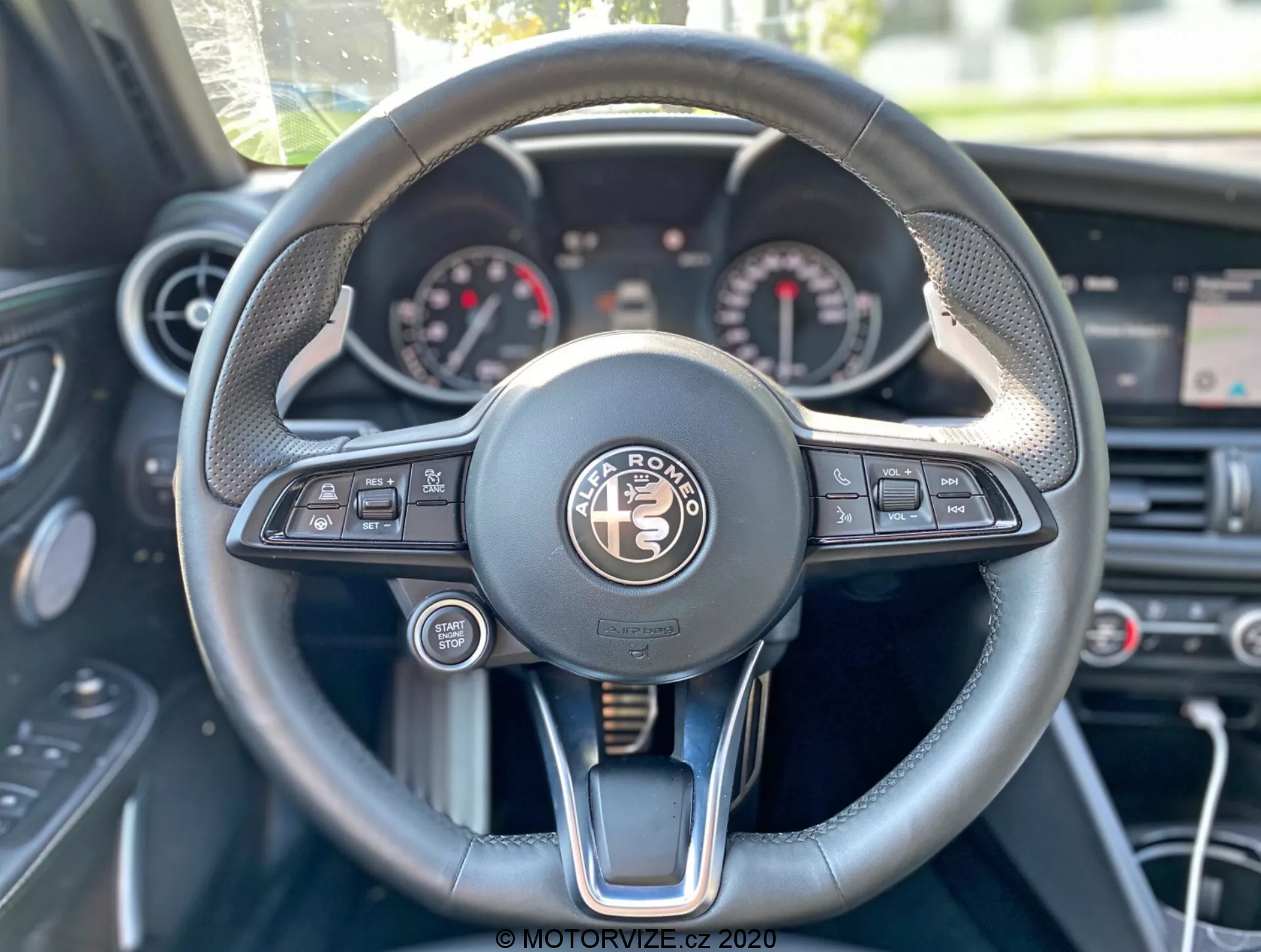 Perspektíva vodiča na interiér Alfa-Romeo Giulia 2016, zameriavajúca sa na volant a palubnú dosku. Centrálne umiestnený volant má multifunkčný dizajn s tlačidlami pre média, hlasové ovládanie, telefónnu konektivitu, informačný displej vozidla a nastavenie tempomatu. Prístrojová doska ukazuje analógové ciferníky, pravdepodobne rýchlomer a otáčkomer, medzi nimi je digitálny displej. Volant kombinuje kožu, šitie a kovové akcenty, vyžarujúci luxus. V pozadí sú viditeľné ovládacie gombíky klimatizácie a nad nimi centrálny displej.