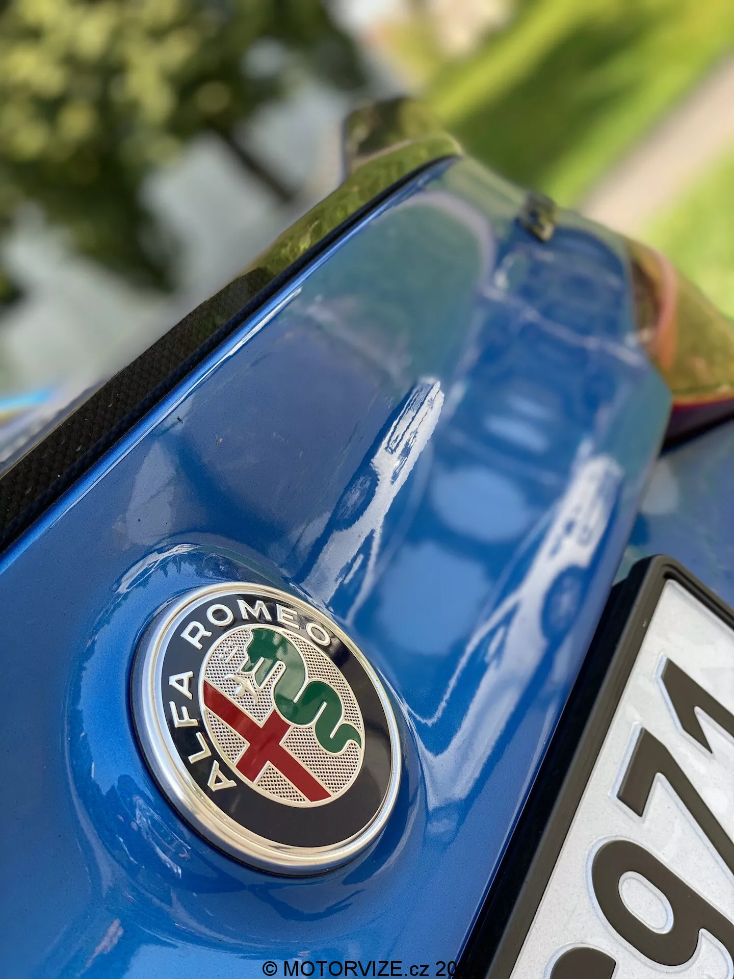 Detailný pohľad na zadnú časť Alfa Romeo Giulia 2016 so zameraním na značkové logo z nízkeho uhla. Odznak s červeným krížom na bielom pozadí a zeleným hadom na modrom pozadí je ostro zaostrený na kovovo modrom veku kufra s detailmi okolitého povrchu a odrazov.