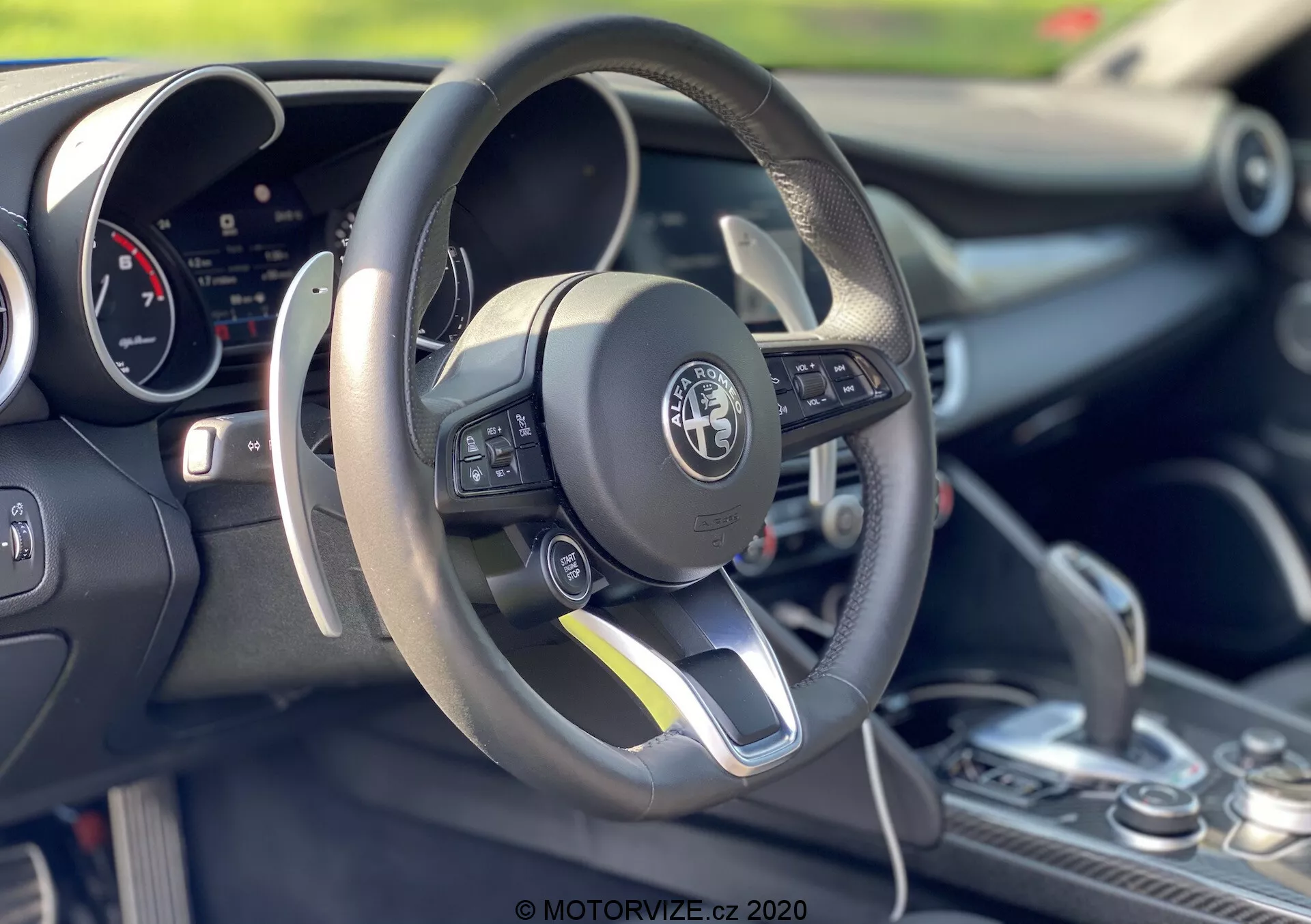 Záber interiéru Alfa-Romeo Giulia z roku 2016 so zameraním na priestor vodiča a volant. Ploský spodný, kožený volant obsahuje multifunkčné ovládače na ľavej strane. Za volantom je viditeľný digitálny prístrojový panel. Stredová konzola ukazuje radiacu páku s koženým prekrytím, čo naznačuje možné manuálne radenie. Obrázok zdôrazňuje športový dizajn, pokročilé technológie a detailné remeselné spracovanie s malou hĺbkou ostrosti.