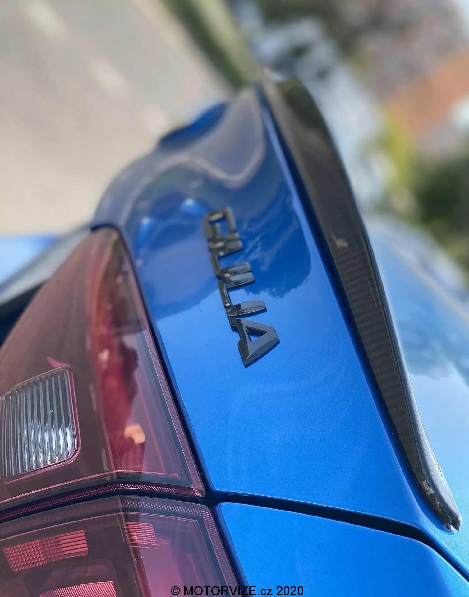 Detailný záber zadnej časti modrého auta ŠKODA z pohľadu zdola hore. Zameriava sa na chrómované označenie 'ŠKODA' na veku kufra, úzky spojler a lesklý, dobre udržiavaný lak. Zadné okno má rozmrazovacie línie a čiastočne je viditeľný moderný pravý zadný svetelný klaster. Nízka hĺbka ostrosti zdôrazňuje značku, pričom predná časť auta a pozadie sú rozmazané. S vodotlačou 'MOTORVIZE.cz 2020', čo naznačuje dátum publikácie alebo vytvorenia.