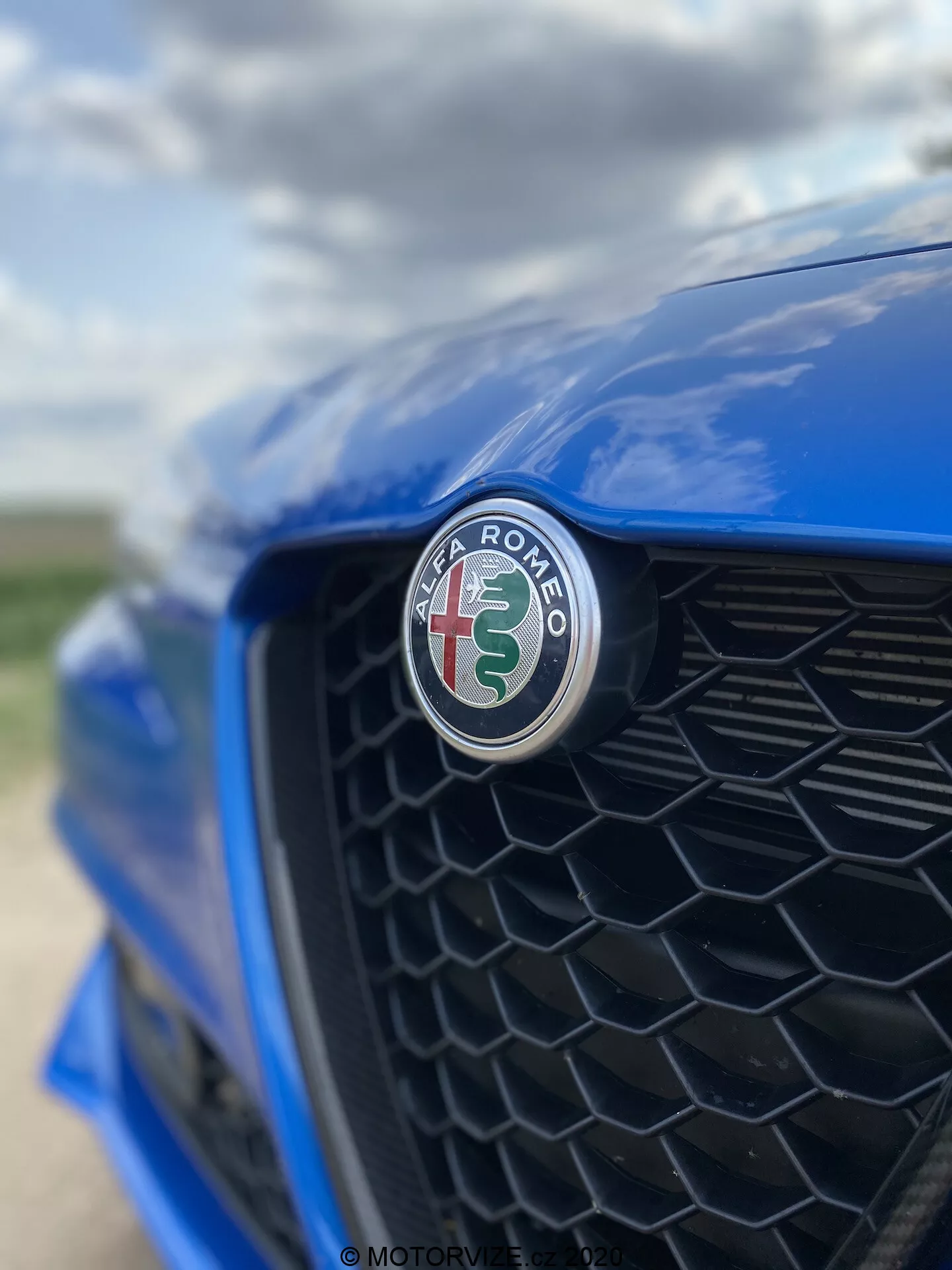 Detailný záber na prednú časť modrého vozidla Alfa Romeo Giulia 2016, zameraný na emblém výrobcu s červeným krížom a zeleným hadom na bielom pozadí. Snímka bola urobená z mimoosového uhla smerom k vodičovej strane, čierna plastová mriežka je jemne rozostrená, čím zvýrazňuje ostré zaostrenie emblému a lesklý povrch.