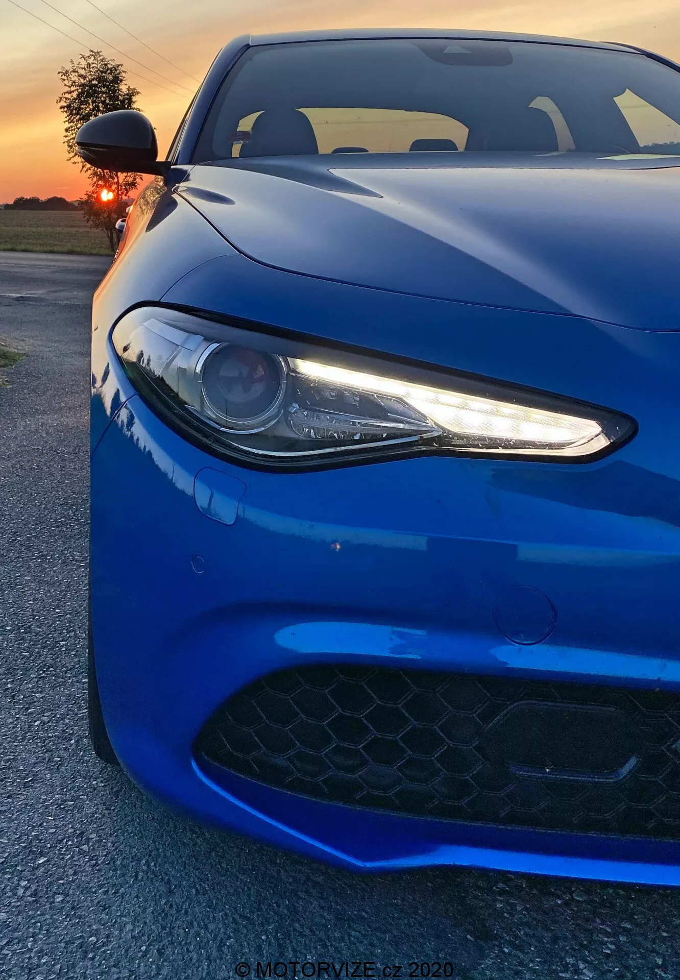 Obrázok zobrazuje športové auto Alfa Romeo Giulia z roku 2016 zachytené z nízkeho predného uhla. Vibrantné hlboké modré vozidlo zapĺňa celý záber, zdôrazňuje jeho ľavý svetlomet s LED dennými svetlami, časť kapoty a mriežku prívodu vzduchu v prednom nárazníku. Šošovkový svetlomet so štíhlymi bielymi LED akcentmi dodáva agresívny, moderný vzhľad. Čierna mriežka so šesťuholníkovým vzorom naznačuje vysokovýkonné schopnosti. Záber zachytáva aerodynamické línie a obrysy auta s odrazy od západu slnka zvýrazňujúcimi jeho lesklú povrchovú úpravu. V pozadí je vidieť teplá oranžová obloha čiastočne zakrytá slnkom. Fotografov blízky uhol zvýrazňuje detaily dizajnu, evokujúc pocit sily a elegancie.