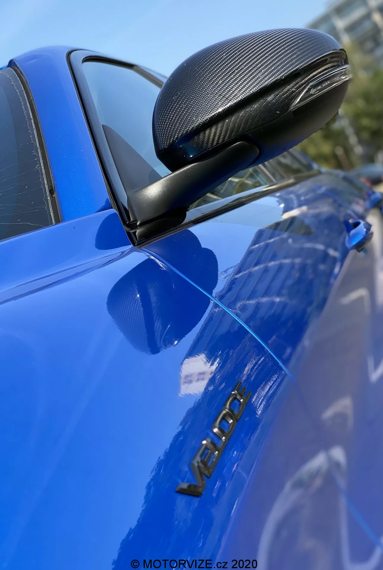 Detailný obrázok modrého auta Alfa Romeo Giulia 2016, zdôrazňujúci spätné zrkadlo s povrchovou úpravou z uhlíkových vlákien v aerodynamickom kryte, namontovanom na čiernej konzole. Fotografia sa ostro zameriava na zrkadlo, vodičove dvere a kovový nápis 'VERON', zvýrazňujúci tieto detaily s malou hĺbkou ostrosti a lesklým, dobre udržiavaným povrchom.