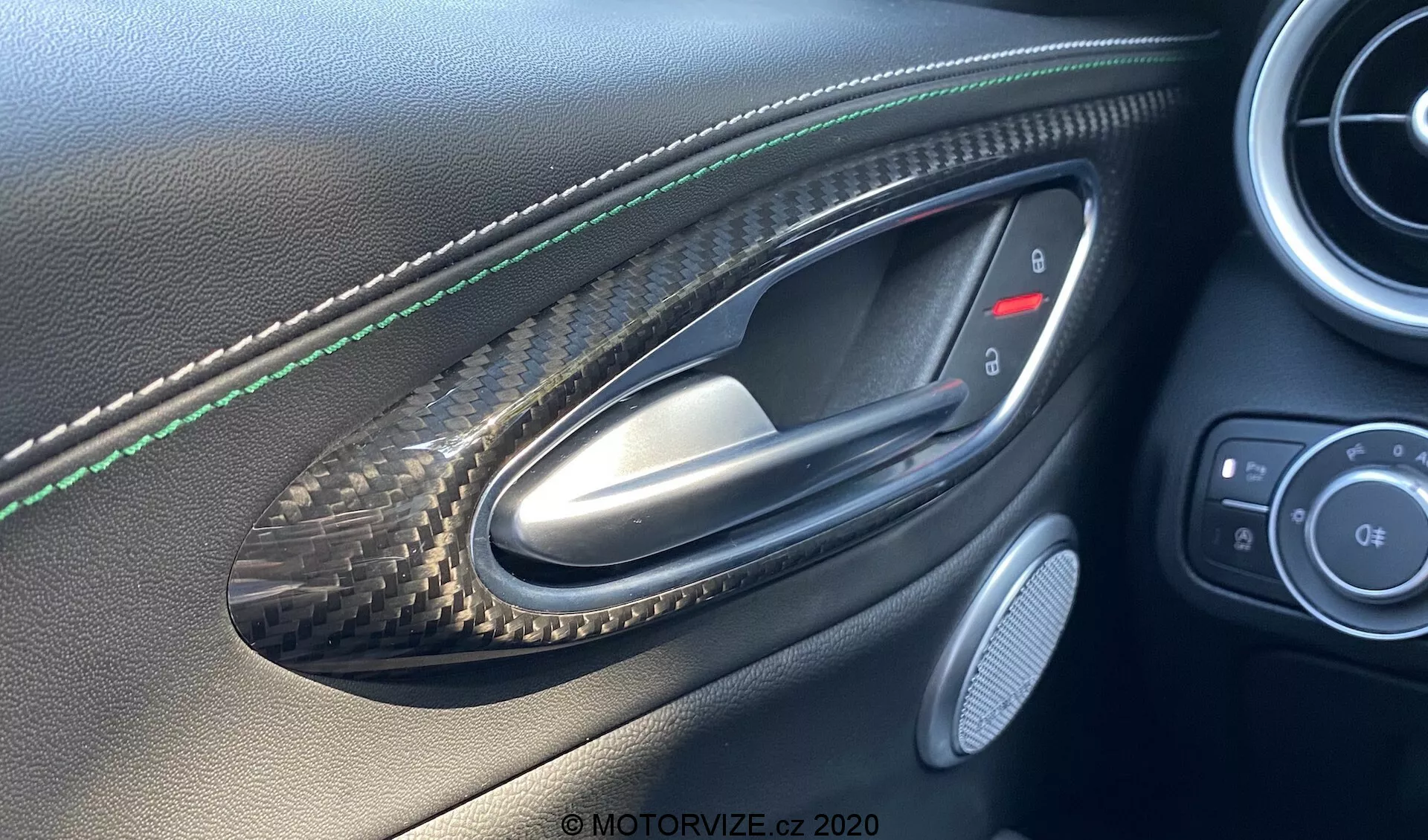 Interiérový záber na dvere vodiča vozidla Alfa Romeo Giulia 2016 z pohľadu človeka sediaceho vo vnútri, zvýrazňujúci kovovú kľučku dverí umiestnenú v prehlbni s karbónovým vzorom, zelené švy na koženom paneli dverí, ovládacie tlačidlá pre spätné zrkadlá a okná, mriežku reproduktora a kvalitné kožené a karbónové lišty, ktoré naznačujú luxusné alebo výkonné vozidlo.