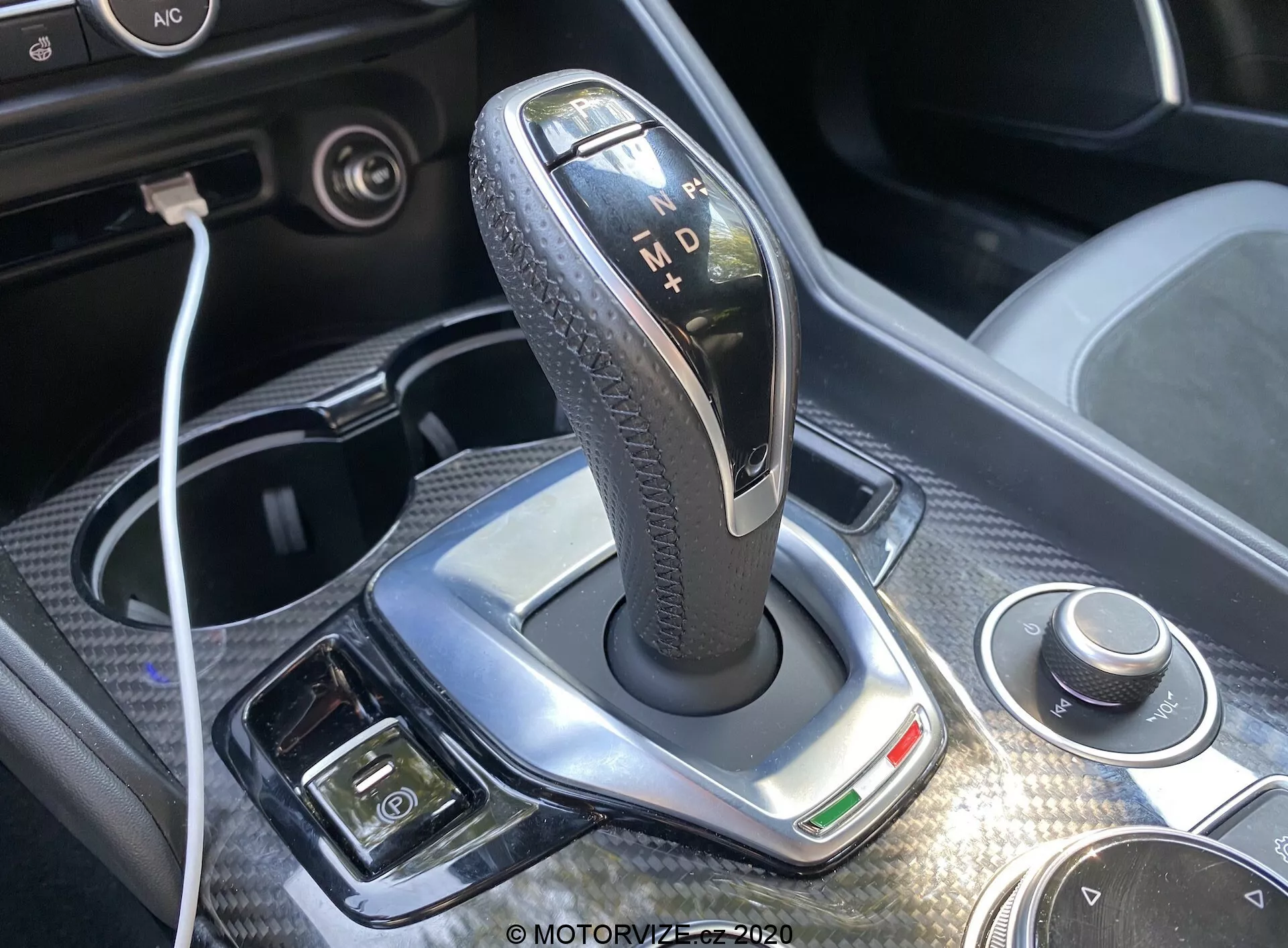 Detailný záber na radiacu páku automatickej prevodovky Alfa-Romeo Giulia 2016 so štýlovým lešteným strieborným okrajom, čiernou koženou textúrou držadla a osvetlenými indikátormi pre 'P', 'R', 'N', 'D' a 'M'. Jasné denné svetlo zvýrazňuje dizajn, pričom v blízkosti sa nachádza otočný volič a pripojiteľnosť USB. Viditeľné je tiež chromované prevedenie, zelený indikačný svetlo a ďalšie tlačidlá pre elektronickú parkovaciu brzdu a klimatizáciu, poskytujúc pohľad na luxus a pokročilé technológie interiéru vozidla.