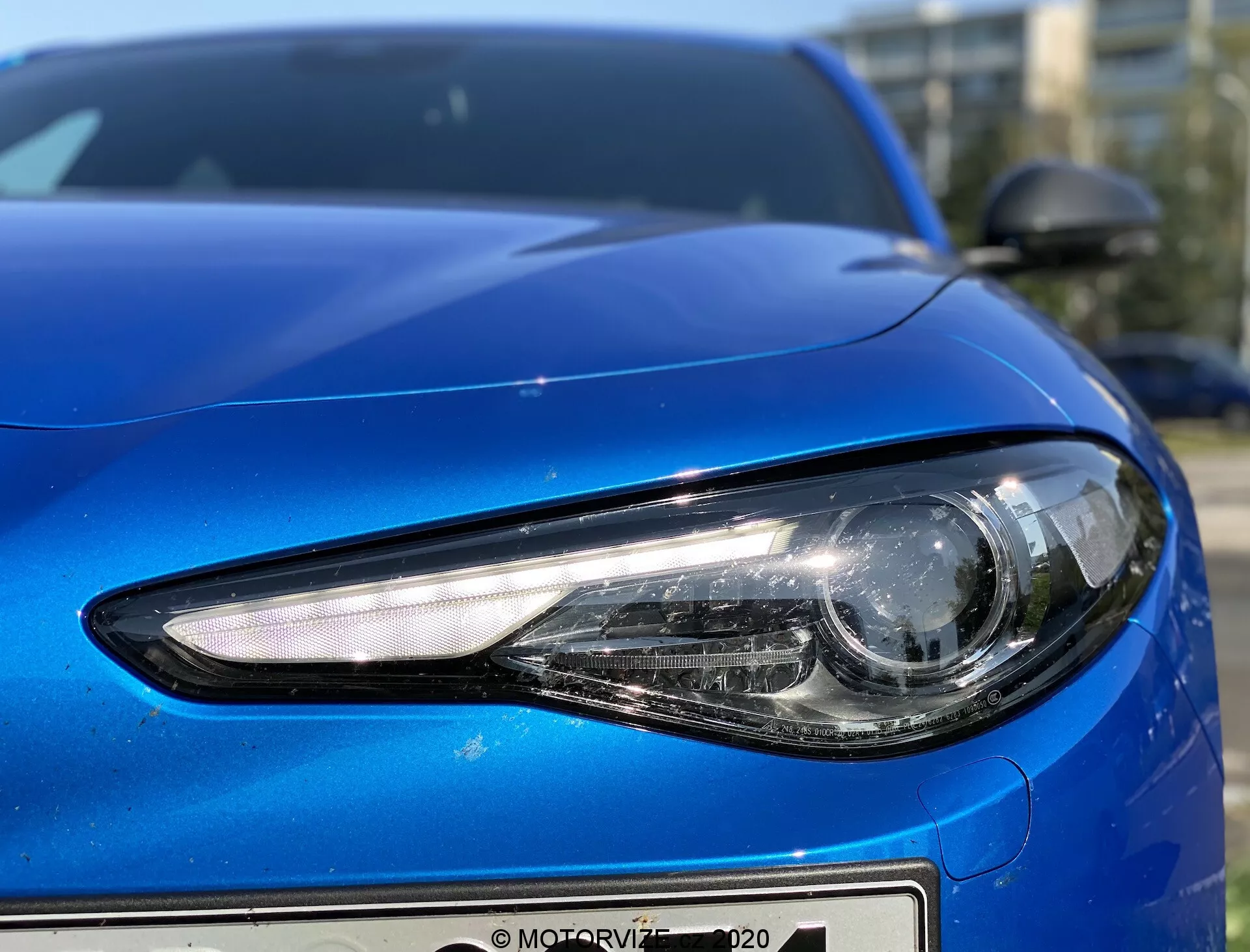 Detailný záber na prednú ľavú stranu modrého auta Alfa Romeo Giulia 2016, zachytený z nízkeho uhla, ktorý ukazuje detaily svetlometu a oblúk dizajnu auta. Moderný svetlomet má LED alebo HID osvetlenie so štíhlym, uhlovým tvarom a projektorovým štýlom hlavného svetla pre zaostrené svetlo. Kapota má hladký, kontúrovaný dizajn s reflexnou kovovou povrchovou úpravou a predný nárazník vykazuje minimalistický dizajn. Zrkadlo v telefarbe je čiastočne viditeľné v pravom hornom rohu a dopĺňa aerodynamický profil auta.