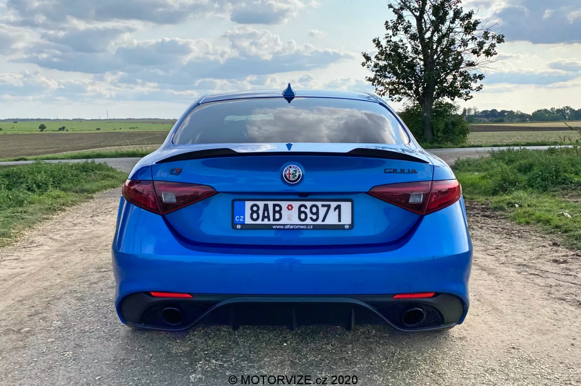 Zadný pohľad na svetlo modrý sedan Alfa Romeo Giulia 2016, zhotovený z úrovne očí. Obrázok zobrazuje celý zadok vozidla, stredne v rámci záberu, s jasne viditeľnou poznávacou značkou, zadnými svetlami a logom výrobcu. Vlastnosti zahŕňajú dvojitý výfukový systém, elegantné moderné zadné svetlá, prvok difuzora na nárazníku, spojler na veku kufra, parkovacie senzory a zadný stierač, čo naznačuje rovnováhu medzi športovým charakterom, bezpečnosťou a praktičnosťou. Nastavenie je vo vidieckej oblasti s plochou krajinou a čiastočne oblačnou dennou oblohou.