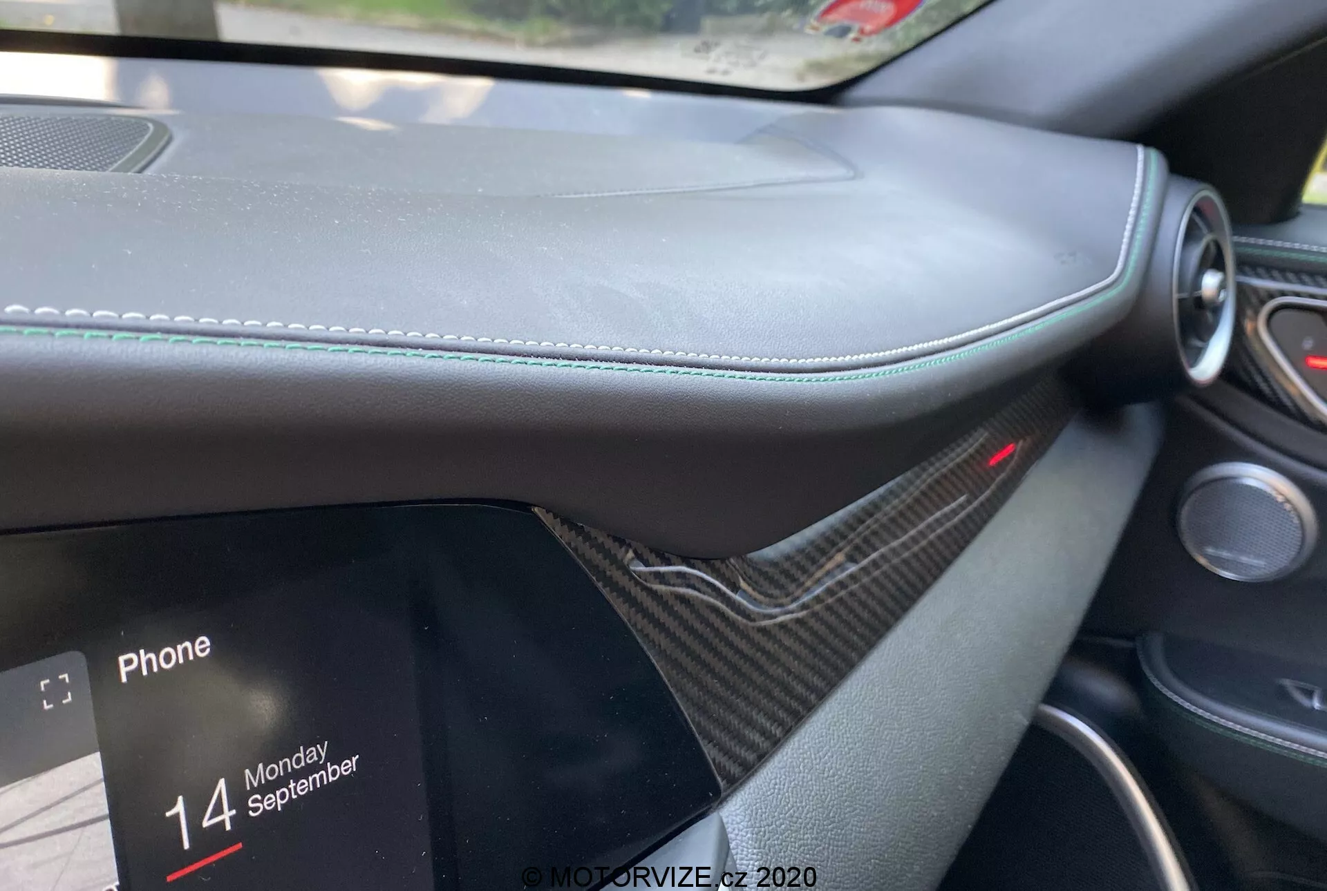 Detail interiéru Alfa Romeo Giulia 2016 z pohľadu spolujazdca, zameraný na palubnú dosku s koženými materiálmi a viditeľným prešívaním, dekórom podobným uhlíkovým vláknam, čiastočne viditeľným infotainment displejom zobrazujúcim možnosti pripojenia telefónu s textom 'Phone' a 'Monday 14 September'. Červené svetlo nad systémom infotainmentu indikujúce aktívny bezpečnostný alebo ochranný systém, zdôrazňujúci luxus a moderný dizajn.