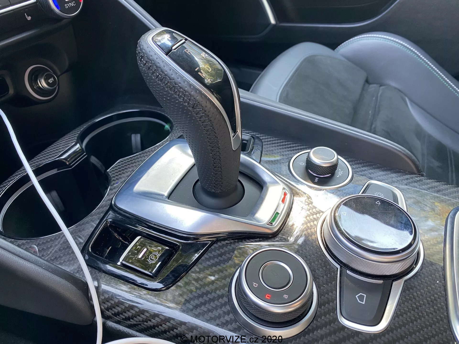 Detailný pohľad na stredovú konzolu a radiacu páku vozidla Alfa Romeo Giulia 2016 s automatickou radiacou pákou potiahnutou kožou a chrómovými akcentmi, obklopenou uhlíkovým zdobením. Konzola obsahuje ovládací gombík typu iDrive, ovládanie hlasitosti, tlačidlá pre dynamiku jazdy, USB port s pripojeným káblom a panel s kontrolkami pohotovostných svetiel a ďalšími ovládacími prvkami, zdôrazňujúc ergonomický a technologicky prepracovaný dizajn interiéru vozidla.