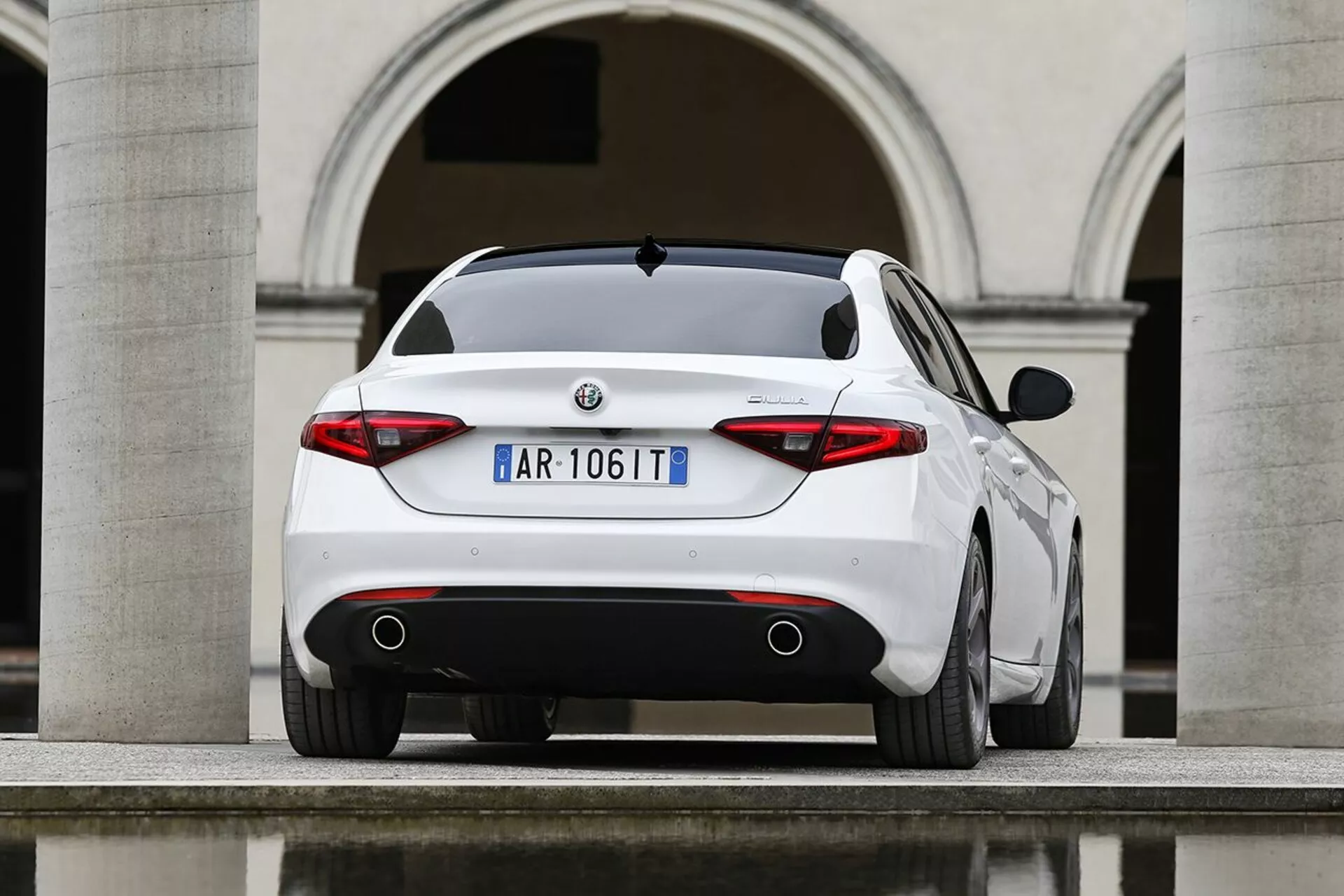 Biela Alfa Romeo Giulia z roku 2016 fotografovaná z uhla zadnej trojštvrtečnej perspektívy, ktorá zdôrazňuje jej športový zadný profil, výrazné zadné svetlá, dvojité výfukové potrubia a štýlové dizajnové prvky na pozadí európskej architektúry