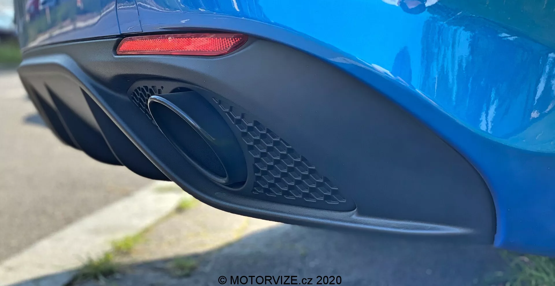 Zblízka bočný pohľad na zadný nárazník a výfukovú časť modrého Alfa Romeo Giulia 2016, ktorý ukazuje jeho moderné dizajnové prvky, vrátane predĺženého horizontálneho červeného LED zadného svetla, veľkého oválneho výfukového otvoru s mriežkou a aerodynamického nárazníka s rôznymi krivkami a preliačinami, fotografovaný s malou hĺbkou ostrosti, ktorá zdôrazňuje detaily auta.