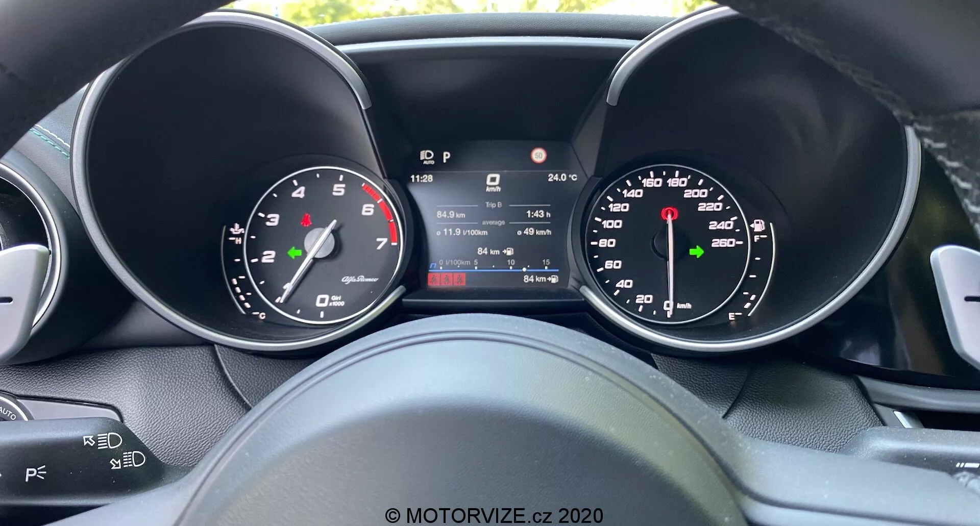 Pohľad vodiča na prístrojovú dosku Alfa Romeo Giulia 2016 s centrálnym digitálnym displejom ukazujúcim rýchlosť, prevodový stupeň, teplotu a stav počítadla kilometrov, po stranách analógového rýchlomera a otáčkomera, predstavujúci kombináciu tradičného dizajnu a modernej technológie s čiastočnými pohľadmi na ovládacie prvky volantu pre audio a tempomat.