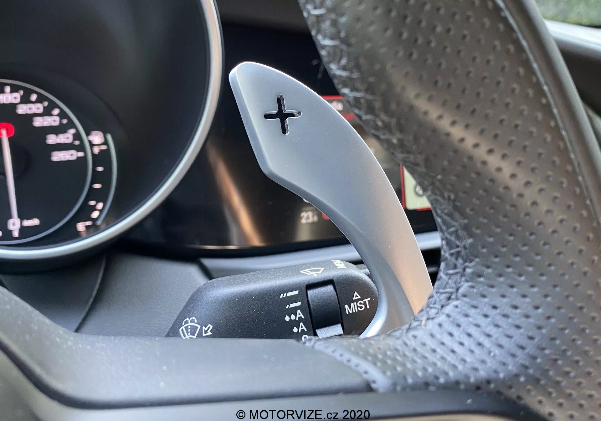 Záber interiéru Alfa Romeo Giulia 2016 so zameraním na pravú radiacu páku s označením '+' na radenie nahor a susednú páku ovládania stieračov na stĺpiku riadenia. Časť otáčkomera z prístrojového panela je mierne viditeľná. Volant z perforovanej kože je mierne rozostrený. Obrázok zvýrazňuje kontrast medzi kovovým radiacim pedálom a matným povrchom páky ovládania stieračov.