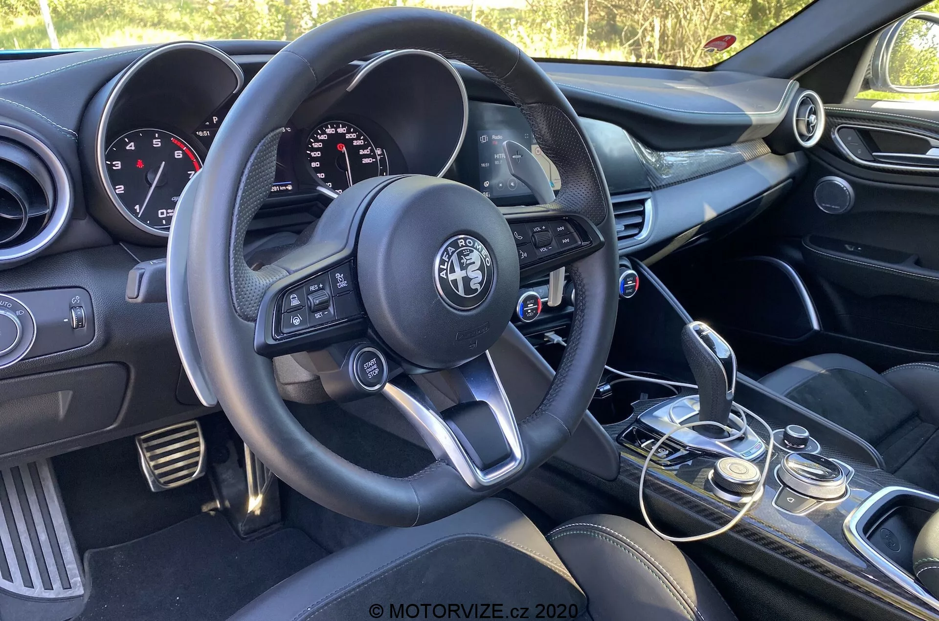 Interiér vozidla Alfa Romeo Giulia 2016 zameraný na stranu vodiča z perspektívy spolujazdca, zobrazujúci trojramenný volant s multifunkčnými tlačidlami, výrazné logo výrobcu, prístrojovú dosku s analógovými ukazovateľmi a digitálnym displejom, centrálnu konzolu s ovládacími tlačidlami audio systému a klimatizácie, manuálnu radiacu páku s koženým obalom, volič jazdných režimov, predné sedadlá s tmavým čalúnením a čiastočne viditeľný ovládací panel dverí vodiča s ovládaním okien a zrkadiel.