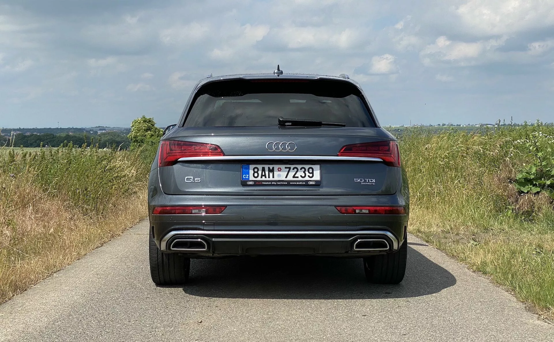 Pohľad zozadu na moderné SUV Audi Q5 2020 Sportback zdôrazňujúci symetriu jeho dizajnu s výraznými LED zadnými svetlami, zadným nárazníkom s kovovými akcentmi a integrovanými dvojitými výfukovými otvormi a chrómovým pruhom nad ŠPZ. Vozidlo stojí na úzkej dlažbe uprostred vidieckej krajiny s poľami a zamračenou oblohou, čím ukazuje svoje luxusné a športové prvky vrátane parkovacích senzorov a strešného spojlera.