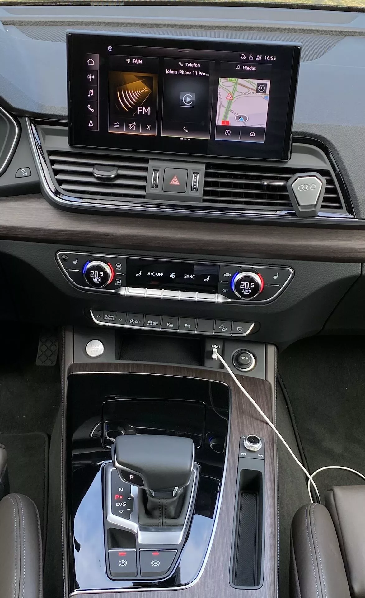 Pohľad zhora na stredovú konzolu a prístrojovú dosku Audi Q5 2020 Sportback, ktorý ukazuje detailné prvky ako dotykovú obrazovku infotainmentu, dvojzónové ovládanie klímy, moderný volič prevodovky s koženým poťahom a luxusným koženým šitím. Perspektíva zdôrazňuje elegantný dizajn, lesklé čierne a kovové akcenty a pokročilé technológie vrátane bezdrôtovej nabíjacej podložky a výberu jazdných režimov.