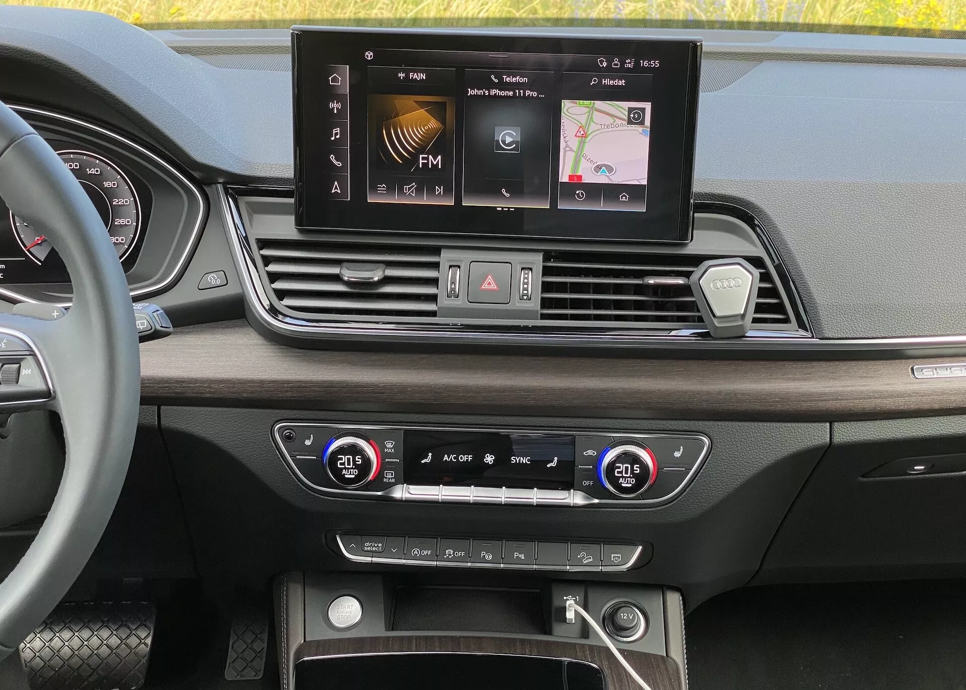 Pohľad na interiérový palubný panel Audi Q5 2020 Sportback z perspektívy očí. Obrázok zvýrazňuje širokouhlý infotainment displej s možnosťami ako FM rádio, navigácia a nastavenie vozidla. Pod ním sú štyri symetricky usporiadané ventilačné otvory, centrálna konfigurácia tlačidiel a gombíkov pre núdzové svetlá, odmrazovanie a rýchly prístup k nastaveniam. Systém dvojzónovej regulácie klímy s digitálnymi displejmi teploty je nastavený na 20,5 stupňa Celzia pre vodiča aj spolujazdca. Spodná časť odhaľuje úložný priestor s možnými USB portmi, pomocným vstupom a tlačidlom start/stop pre technológiu bezkľúčového zapaľovania. Luxusné materiály a dizajn zahŕňajú drevené dekorácie a leštené kovové akcenty.