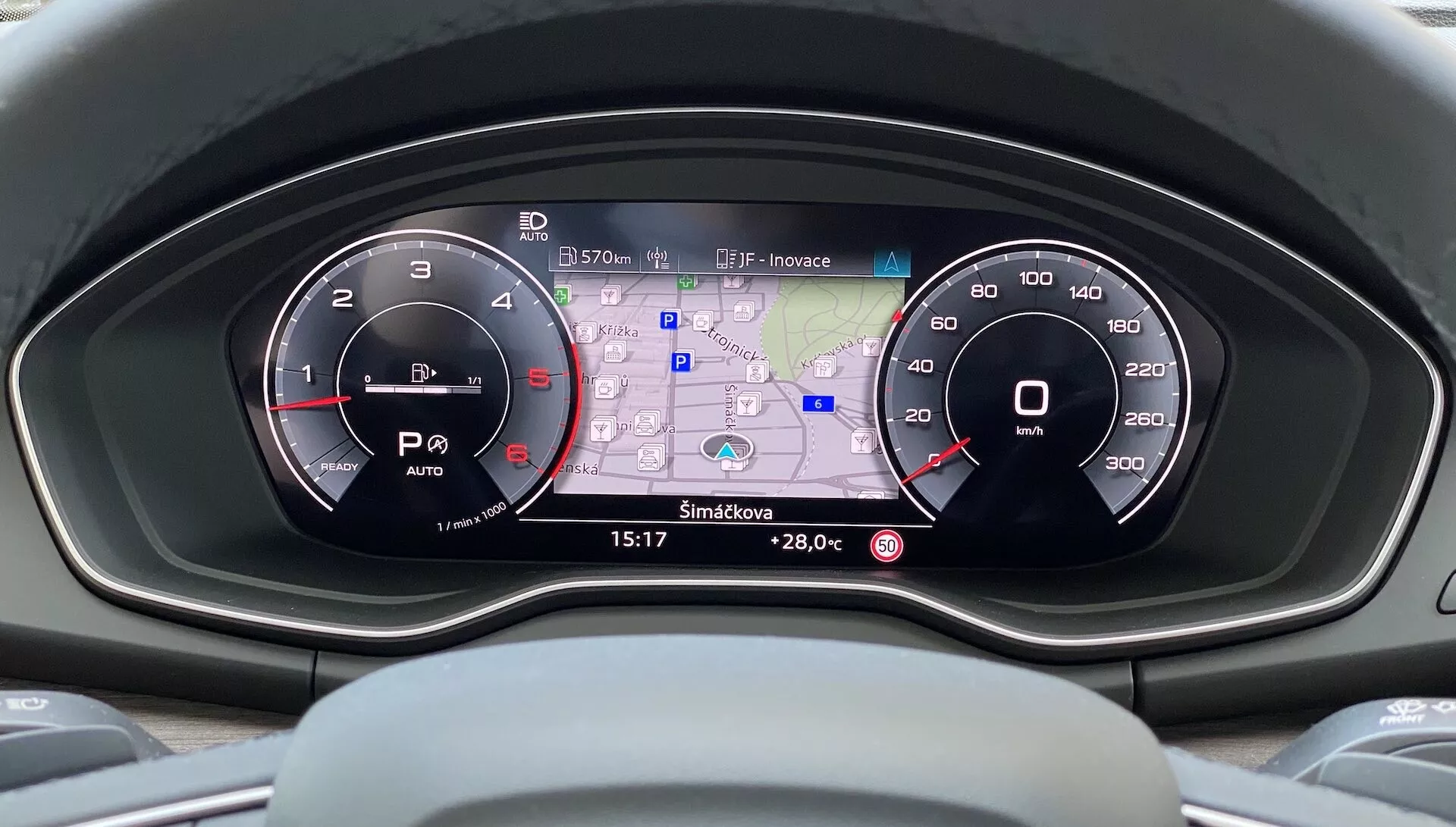 Pohľad od vodiča na moderný prístrojový panel Audi Q5 Sportback 2020, ktorý ukazuje prístrojový panel s dvoma analógovými číselníkmi a centrálnym digitálnym displejom s GPS mapou, údajom o teplote, spotrebe paliva, indikátormi jazdných režimov a časom na vysokorozlíšenej farebnej obrazovke.