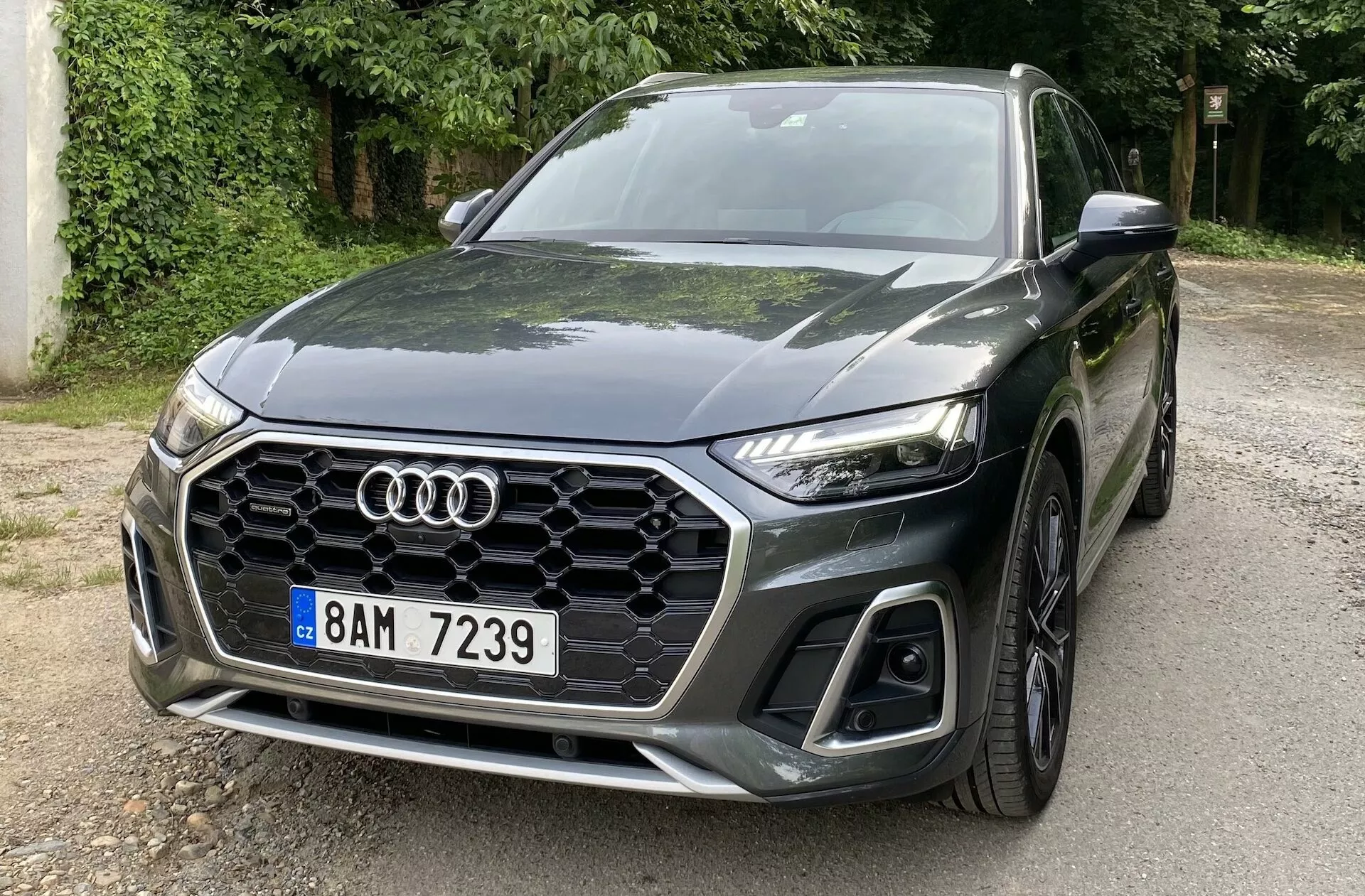 Trojštvrťový pohľad spredu na Audi Q5 Sportback 2020, ktorý zobrazuje ľavú prednú stranu s charakteristickou šesťuholníkovou mriežkou s medovým vzorom, logom Audi, elegantnými hranatými LED svetlami a veľkými nasávacími otvormi na spodnom nárazníku. Auto je zaparkované na ceste obklopenej stromami, čo odráža jeho tmavú, lesklú farbu. Vyrezávaná kapota s aerodynamickými líniami smerujúcimi k emblému a predné koleso s viaclúčovým dizajnom zdôrazňujú jeho športový, moderný dizajn.