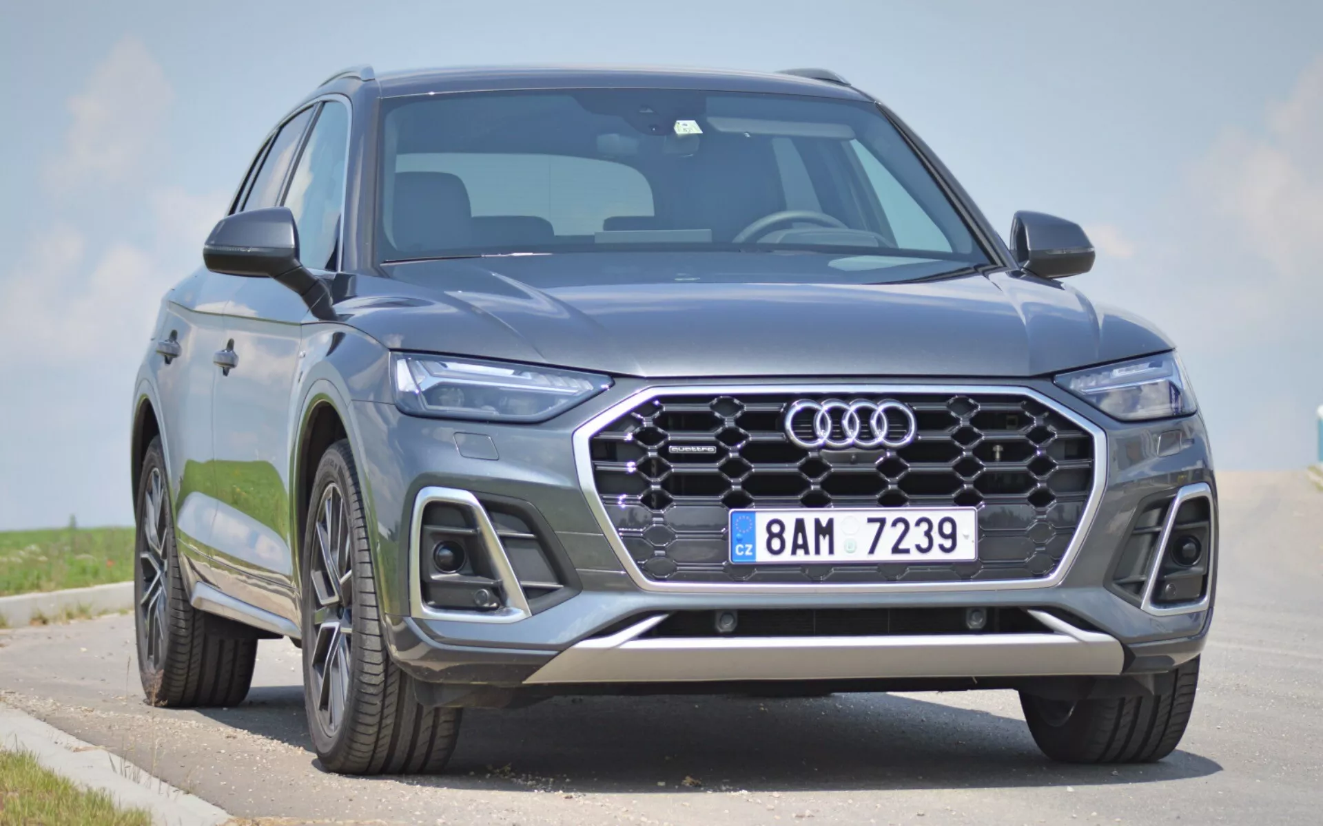 Moderné Audi Q5 2020 (Q5 Sportback) SUV zachytené z nízkeho predného trojštvrťového uhla, zobrazujúce prednú fasádu, šesťhrannú mriežku s emblémom štyroch krúžkov, elegantné LED svetlomety a časť bočného profilu. Veľká vzorovaná mriežka, integrované hmlové svetlá, nasávacie otvory a strieborné ozdobné prvky na prednom nárazníku pridávajú estetický a funkčný dojem. Pokročilé osvetľovacie a asistenčné technológie pre vodiča naznačuje ostrý dizajn svetlometov a konfigurácia mriežky. Veľké zliatinové kolesá a dynamické línie karosérie naznačujú prémiovú, robustnú prítomnosť. Nepohyblivé vozidlo na ceste s čistou oblohou na pozadí zdôrazňujú predné prvky a agresívne štýlové prvky vozidla.