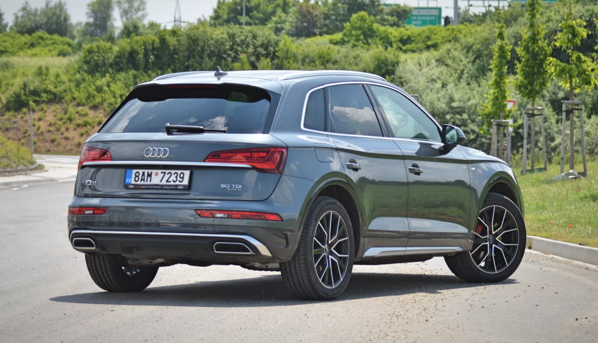 Obrázok zobrazuje moderné Audi Q5 2020 Sportback SUV zaparkované na vydláždenej ploche za denného svetla, s trojštvrťovým zadným uhlom pohľadu. Na zadnej časti vozidla vidno logo Audi a označenie modelu, s elegantnými horizontálnymi zadnými svetlami, ktoré plynulo prechádzajú do bočného profilu. Lesklá kovová tmavozelená farba vozidla odráža svetlo a zobrazuje jeho kontúry. Bočné okná na strane spolujazdca majú tónované sklá, čo naznačuje prémiovú kategóriu. Jemne klesajúca línia strechy dodáva športový vzhľad, akcentovaný strešným spojlerom. Zliatinové kolesá s viacerými lúčmi a viditeľné kotúčové brzdy naznačujú vyššiu úroveň výbavy. Svetlá výška SUV a ochranné obloženie karosérie pozdĺž bočných prahov a okolo blatníkov naznačujú univerzálnosť a schopnosť jazdy v teréne. Strieborné strešné nosníky naznačujú ďalšiu funkčnosť a kľučky dverí a bočné zrkadlá sú vo farbe karosérie. Dvojité výfuky v zadnom nárazníku naznačujú výkonný motor.