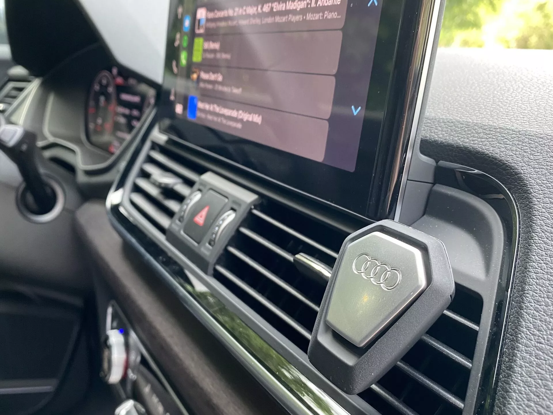 Detailný pohľad zo strany vodiča na centrálnu palubnú dosku Audi Q5 Sportback 2020. Zameranie je na centrálny ventilačný otvor a infotainment displej, ktorý zobrazuje prehrávanie médií so zoznamom zvukových súborov, čím zdôrazňuje multimediálne schopnosti vozidla. Ventilačné otvory sú obdĺžnikového tvaru s kovovým povrchom a obsahujú gombíky s logom Audi. Palubná doska má textúrovaný povrch prémiovej kvality, čo je v súlade s luxusným umiestnením značky Audi, a chýba viditeľné manuálne ovládanie klimatizácie, čo naznačuje automatický klimatizačný systém.