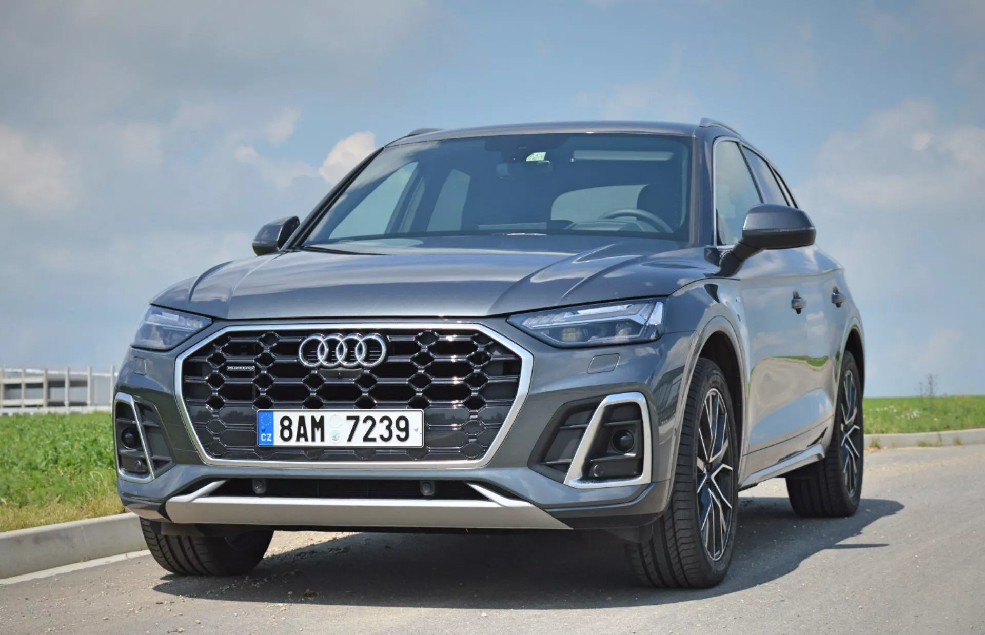Obrázok zobrazuje moderné SUV Audi Q5 Sportback z roku 2020 zachytené z nízkeho a blízkeho uhla, zvýrazňujúc prednú časť vozidla a masku chladiča. Výrazné črty zahŕňajú ikonické štyri kruhy Audi, charakteristickú mriežku s dizajnom plástov, elegantné LED svetlomety s charakteristickými dennými svetlami, integrované snímače v prednom nárazníku indikujúce pokročilé asistenčné systémy vodiča, veľké viaclúčové kolesá z ľahkej zliatiny, ostré línie karosérie a aerodynamické bočné zrkadlá. Vozidlo je zaparkované pod čistou oblohou, zdôrazňujúc jeho luxusný a technologicky pokročilý dizajn.