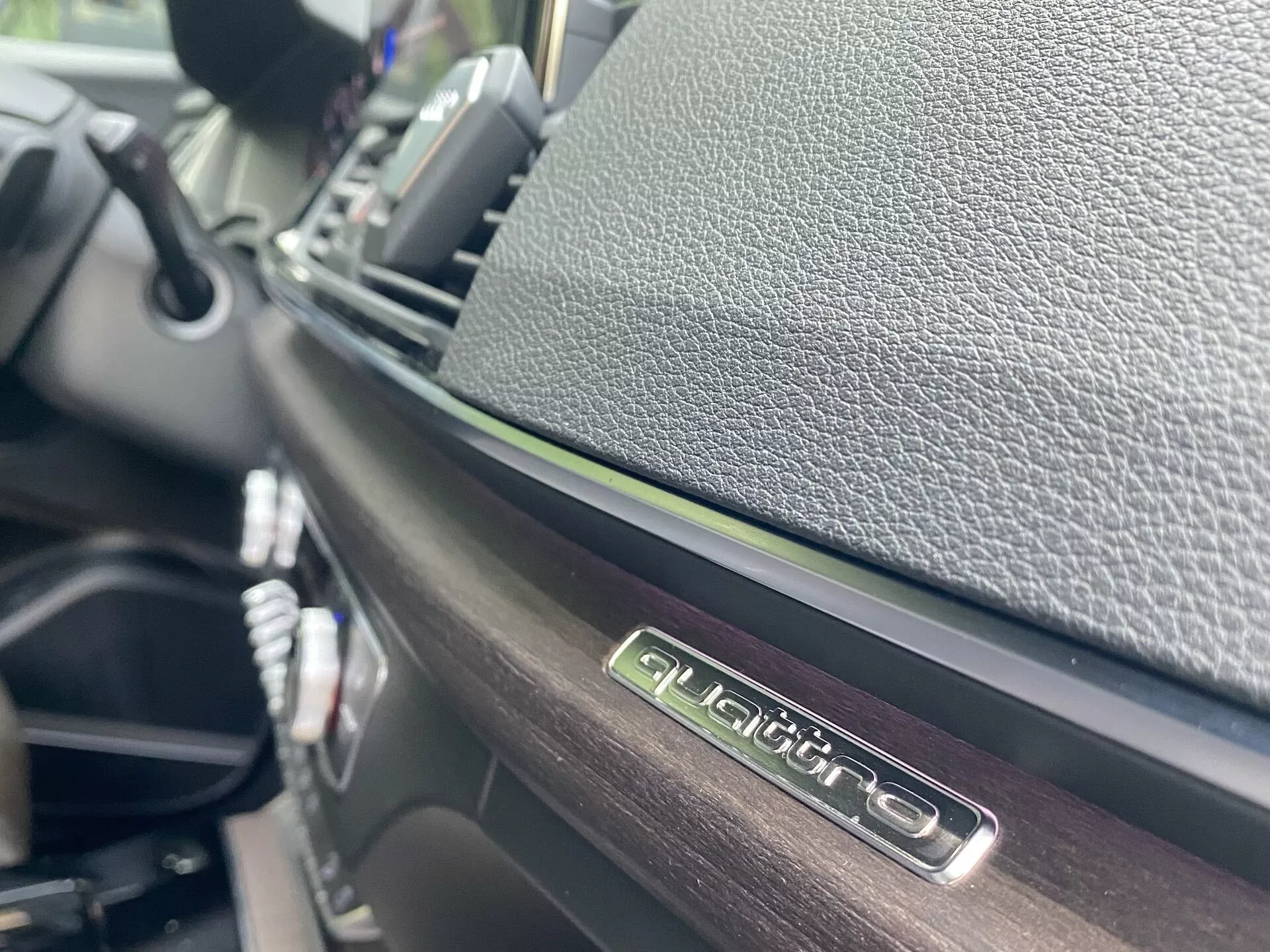 Detailný pohľad na interiér Audi Q5 2020 Sportback so zameraním na znak 'QUATTRO' na úrovni očí, ktorý zdôrazňuje pohon všetkých štyroch kolies Audi. Lesklý povrch znaku a kontrastné texturované materiály, urobené z uhlového uhla s nízkou hĺbkou ostrosti, ukazujú detaily palubnej dosky a ovládacieho panela v rozmazanom pozadí.