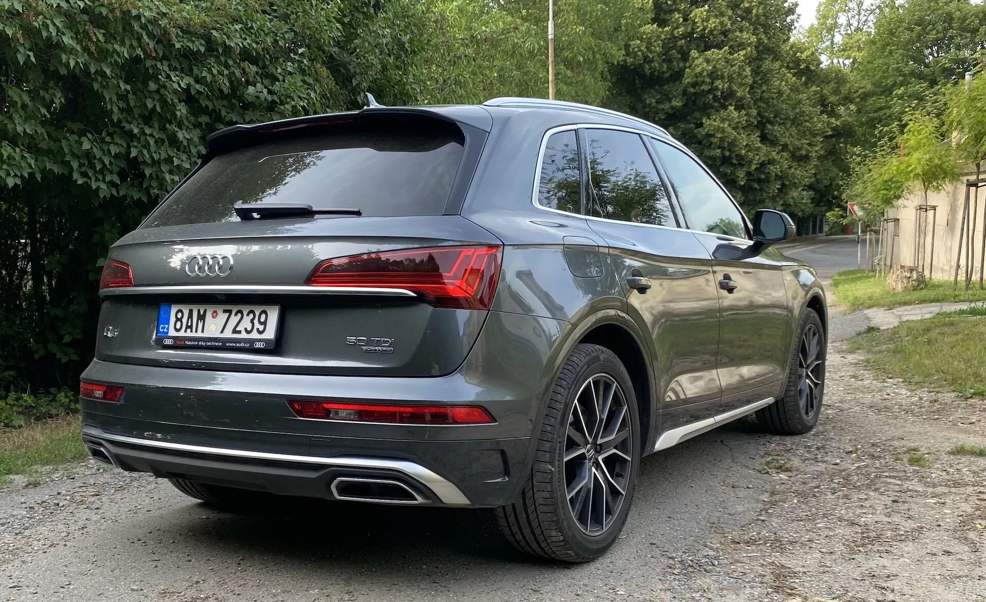 Zadný uhol pohľadu na sivé Audi Q5 Sportback 2020 SUV zaparkované na štrkovom povrchu vedľa zelenej vegetácii. Obrázok sa sústreďuje na zadný a bočný profil vozidla, ukazujúc moderné LED zadné svetlá, dvojité výfukové potrubia, logo Audi kruhy, znaky modelu 'Q7' a variantu '45 TDI', viacramenné hliníkové kolesá, zatmavené sklá, strešný spojler a potenciálne parkovacie senzory alebo kameru. Prirodzené osvetlenie s mäkkými tieňmi naznačuje východ alebo západ slnka.