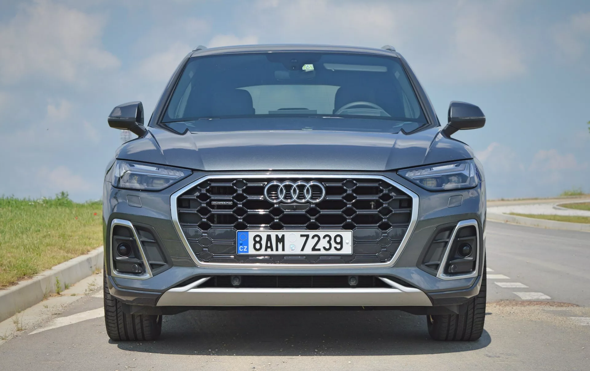 Čelný pohľad na moderné Audi Q5 2020 Sportback SUV, ktorý ukazuje jeho elegantnú a symetrickú prednú časť s ikonickou mriežkou Audi, LED svetlometmi, parkovacími senzormi, kovovým modrým lakovaním, zliatinovými kolesami a strieborným obložením. Scéna je zasadená na dláždenej ceste pod jasnou oblohou.