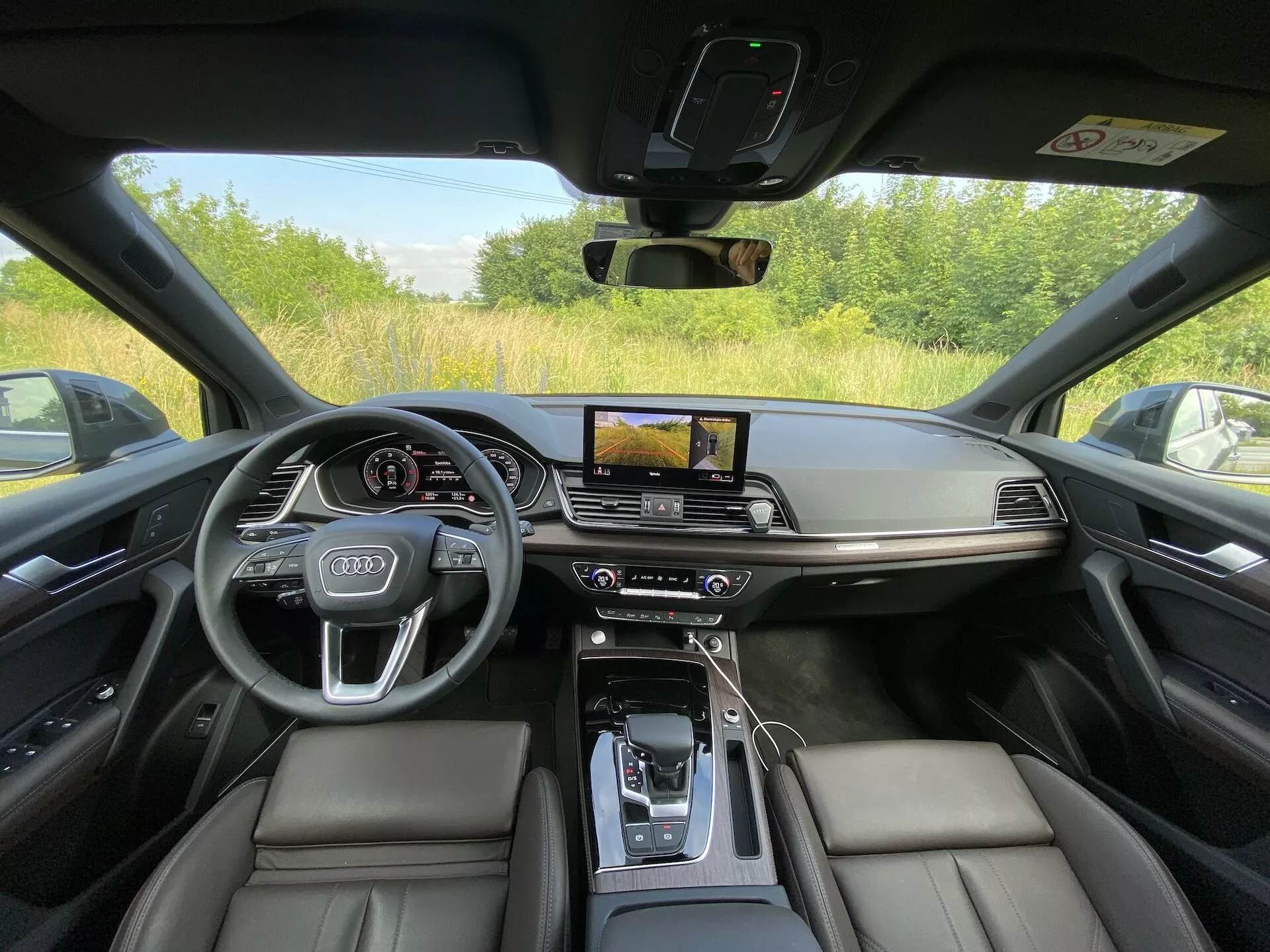 Interiér Audi Q5 Sportback 2020 odfotený z perspektívy sedadla vodiča, ktorý ukazuje luxusný a technologicky vyspelý prístrojový panel. Obrázok zdôrazňuje čierne kožené sedadlá, stredovú konzolu s koženou opierkou na ruku, automatickú radiacu páku obklopenú ovládacími tlačidlami, vodorovne orientovaný dotykový displej infotainmentu s GPS navigáciou, dvojzónovú klimatizáciu, digitálny displej vodiča za volantom s logom Audi a namontované ovládacie prvky. Dizajn sa vyznačuje minimalizmom, kovovými akcentmi a úplnou viditeľnosťou cez okno a stĺpik vozidla, čo naznačuje, že je zaparkované vonku.