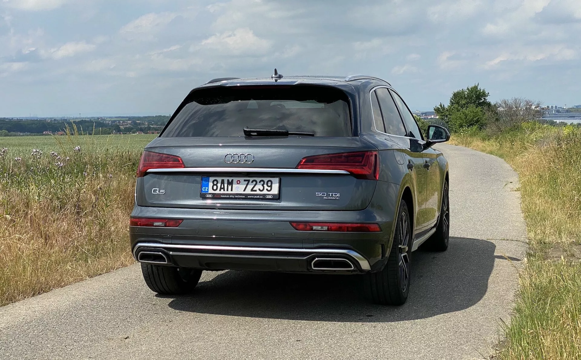 Zadný pohľad na Audi Q5 Sportback z roku 2020, zobrazujúci šírku vozidla vrátane zadných svetiel, zadného skla a dvojitých výfukových rúrok. Auto, umiestnené na ceste za denného svetla, ukazuje vidiecke okolie a jeho lesklý lak odráža okolitú krajinu. Sú zreteľne viditeľné detaily dizajnu ako logo Audi, označenie modelu a evidenčné číslo. Technologické prvky, ako sú LED svetlá, parkovacie senzory a odmrazovanie zadného skla, sú tiež zrejmé.