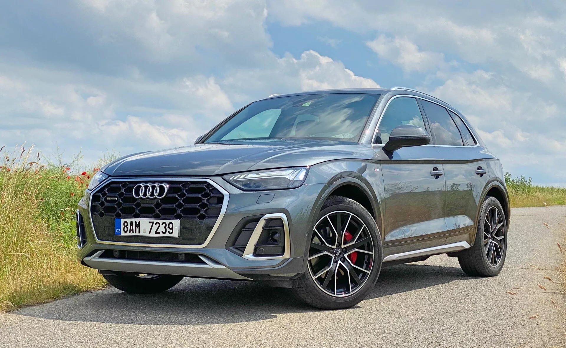 Obrázok zobrazuje moderné SUV Audi Q5 Sportback 2020 zaparkované na dláždenej bočnej ceste v prírodnom prostredí, pravdepodobne v vidieckej oblasti. Výrazné v popredí je logo Audi so štyrmi kruhmi. Tento detailný záber z úhlu troch štvrtín zvýrazňuje prednú ľavú stranu a čiastočný profil vozidla, zdôrazňujúc dizajnové prvky ako šesťuholníkovú mriežku na prednej maske, ostré LED svetlomety a veľké viacpaprskové kolesá s čiernymi detailmi a červenými brzdovými strmeňmi. Kovová šedá farba odráža svetlo a zdôrazňuje kontúry vozidla. Mierne zvýšená svetlá výška SUV naznačuje univerzálnosť a ľahké schopnosti v teréne, zatiaľ čo bočné zrkadlá zobrazujú kontrastnú farbu, ktorá naznačuje športovú verziu. Obrázok stelesňuje luxusný a športový automobilový dizajn s potenciálnymi pokročilými technológiami, ktoré nie sú viditeľné.