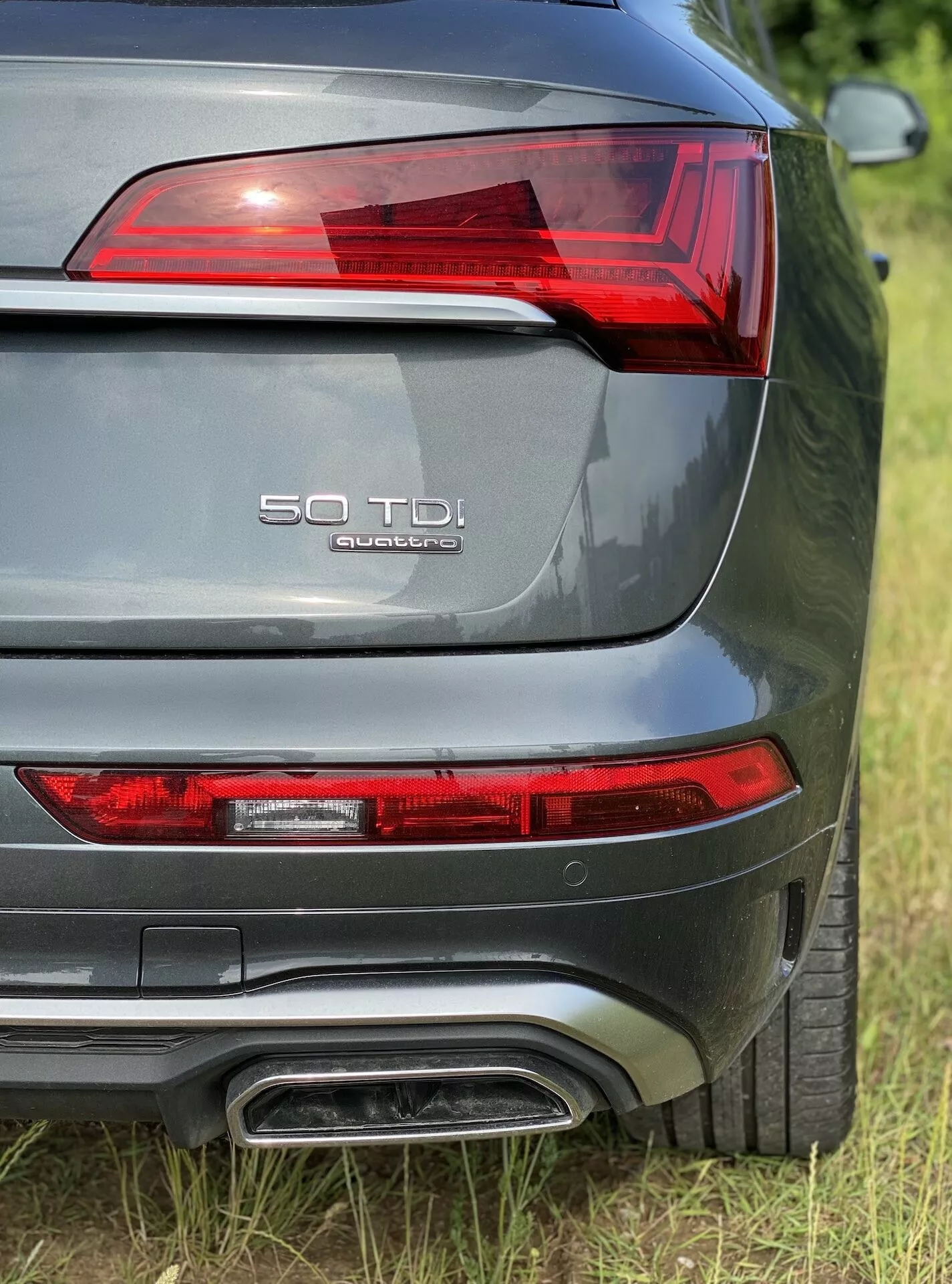 Detailný pohľad na zadnú časť Audi Q5 2020 Sportback z trojštvrťového zadného pohľadu, zobrazujúci ľavé zadné svetlo s LED prvkami, označenie modelu '50 TDI' a 'quattro' na veku batožinového priestoru, dvojitý výfukový systém, kovovo sivú farbu a viditeľný zadný parkovací senzor.