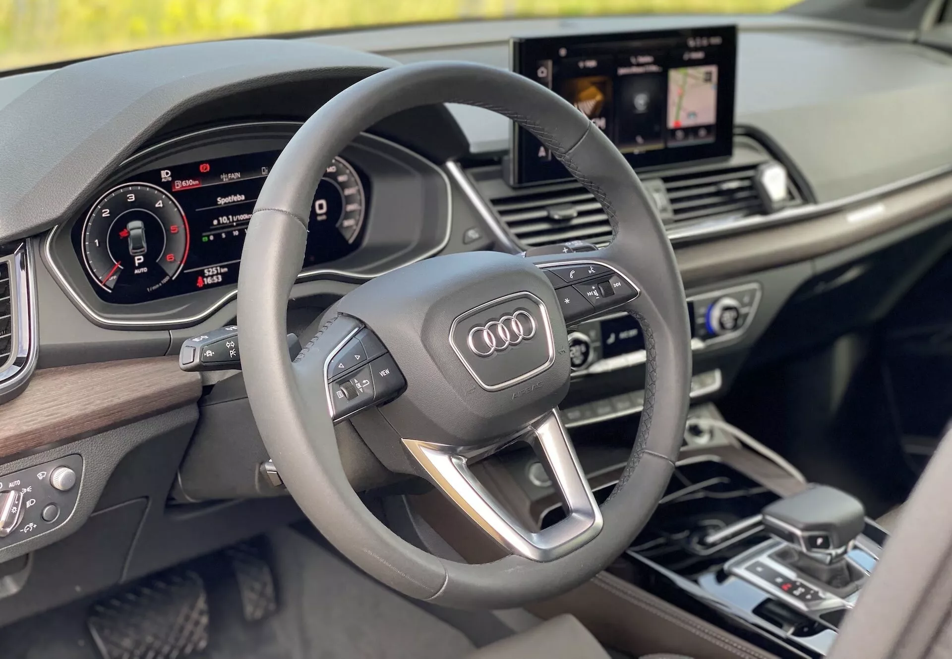 Luxusný interiér Audi Q5 2020 Sportback z pohľadu vodiča, predstavujúci moderný trojramenný multifunkčný volant s integrovanými ovládačmi, plne digitálny prístrojový panel a centrálne umiestnenú informačnú obrazovku zobrazujúcu navigáciu. Interiér je vybavený drevenými ozdobnými prvkami, ktoré zvýrazňujú pocit luxusu a modernosti.