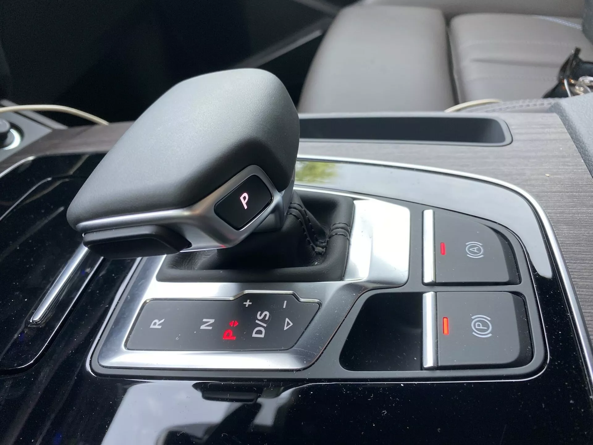 Detailný pohľad na automatickú prevodovku Audi Q5 2020 Sportback v stredovej konzole, s dizajnom shift-by-wire v parkovacej polohe 'P', s možnosťami 'R' (spätný chod), 'N' (neutrál) a 'D/S' (jazdný/sportový režim), spolu so znakmi '+' a '-' pre manuálny posun, tlačidlom 'Auto Hold' a elektronickou ručnou brzdou. Okolo prevodovky sa nachádza lesklá čierna úprava a chrómové akcenty naznačujúce luxusný a technologicky pokročilý interiér.