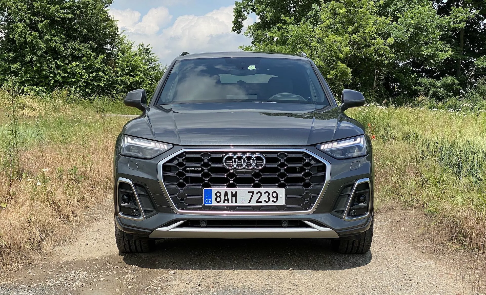 Predný pohľad na SUV Audi Q5 Sportback 2020 so silnou šesťuholníkovou maskou Singleframe s vertikálnymi chrómovými prúžkami, centrálnym umiestnením znaku Audi, ostrými Matrix LED svetlometmi s charakteristickými dennými bežnými svetlami, viditeľnými prieduchmi so vzorovaným dizajnom a predným nárazníkom s integrovanými hmlovými svetlami a kontúrovanými líniami. Auto je postavené na štrkom alebo zeminovým povrchu, v prírodnom vidieckom prostredí s denným svetlom vrhajúcim tieňe, čo zdôrazňuje elegantný športový vzhľad a pokročilé technologické prvky.