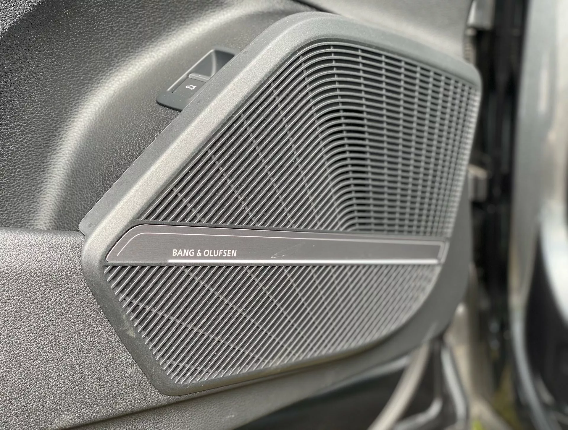 Detailný záber na prémiovú mriežku reproduktora BANG & OLUFSEN na dverovom paneli Audi Q5 Sportback 2020. Jemný vzor mriežky je ostro zaostrený, zatiaľ čo pozadie a popredie sú rozmazané. Nad mriežkou je viditeľná malá trojuholníková časť, ktorá môže obsahovať výškový reproduktor, z bočného a mierne predného uhla. Obraz zdôrazňuje luxusnú kvalitu zvukového systému vozidla.